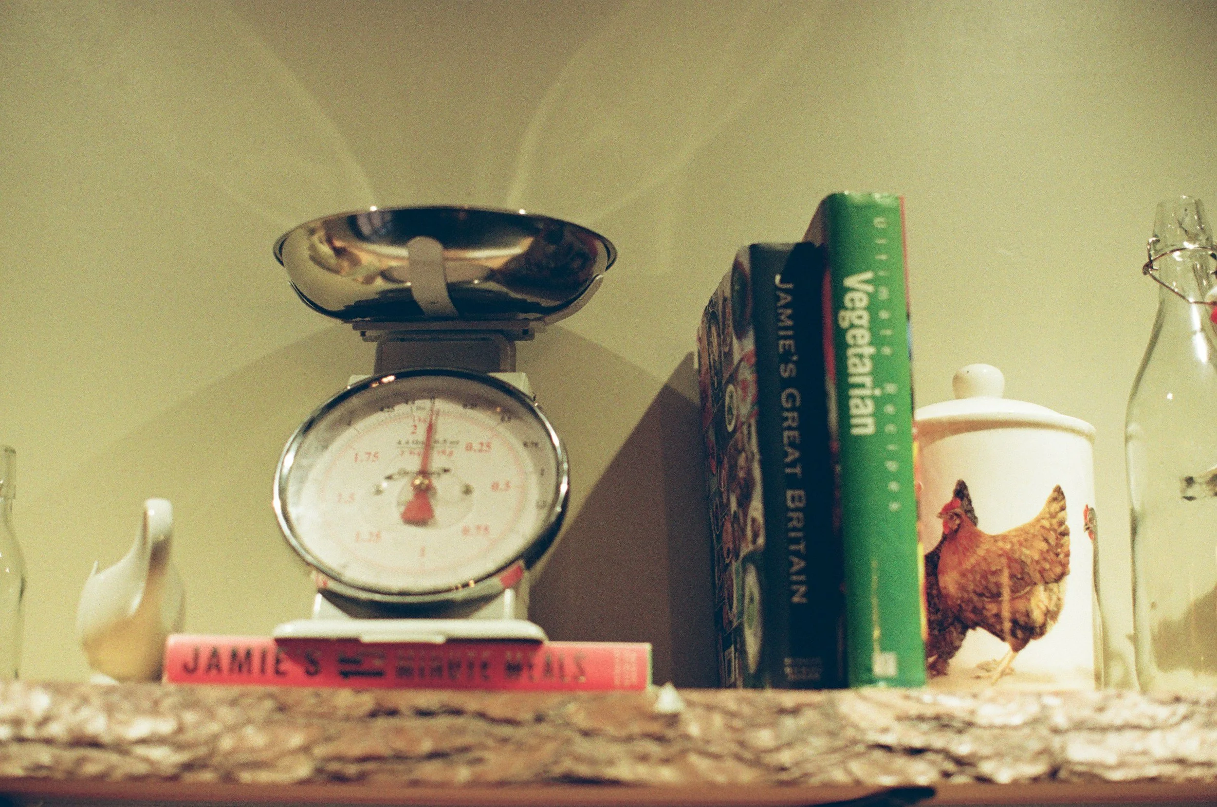 Kitchen counter with a vintage kitchen scale, stacked books including 'Jamie’s' and some board games, a ceramic chicken figurine, a milk glass vase, and a cup with a spoon inside.