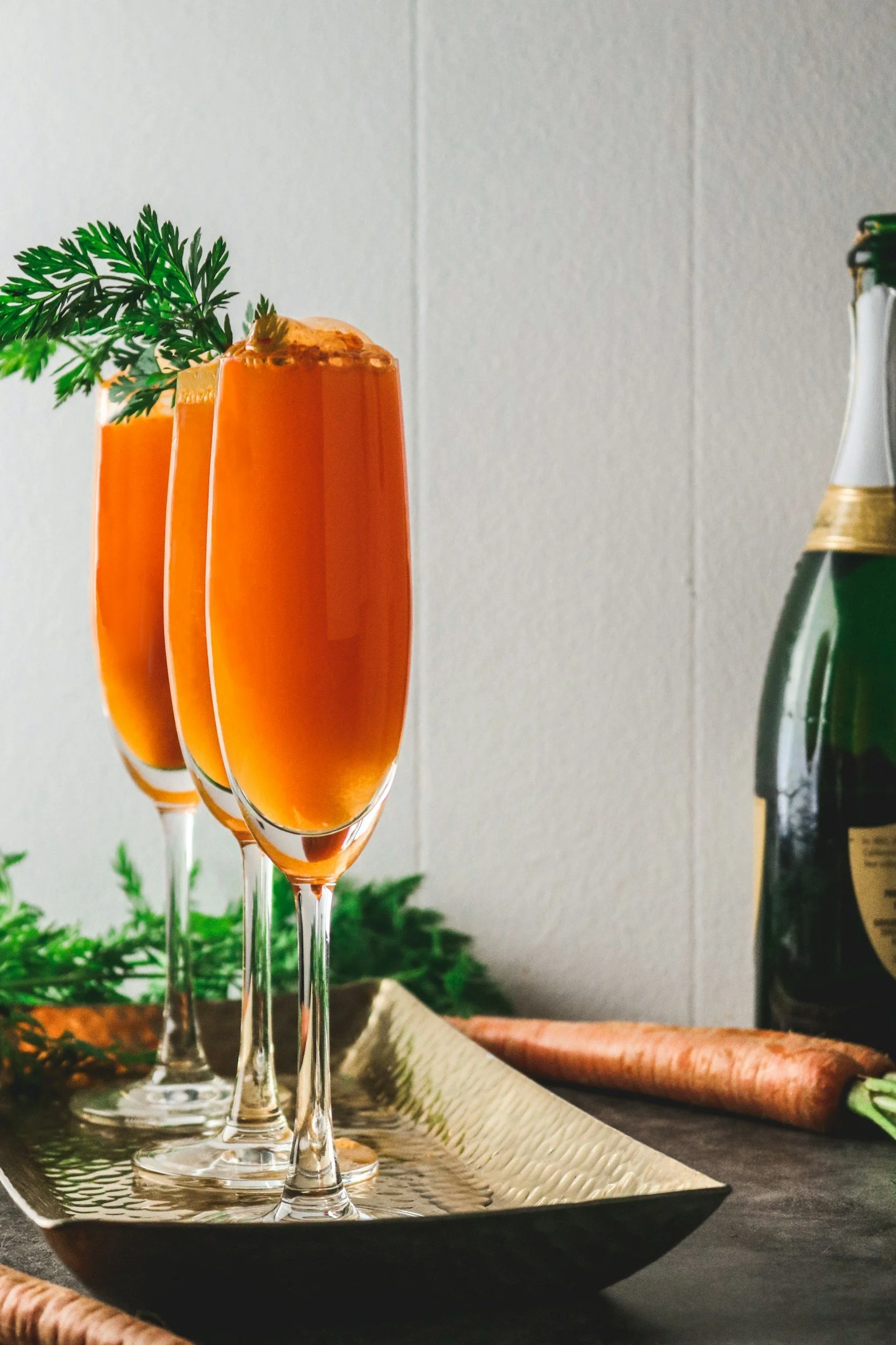 Three filled champagne glasses with orange beverage, garnished with a green sprig, placed on a silver tray with a textured pattern, surrounded by greenery and a champagne bottle in the background.