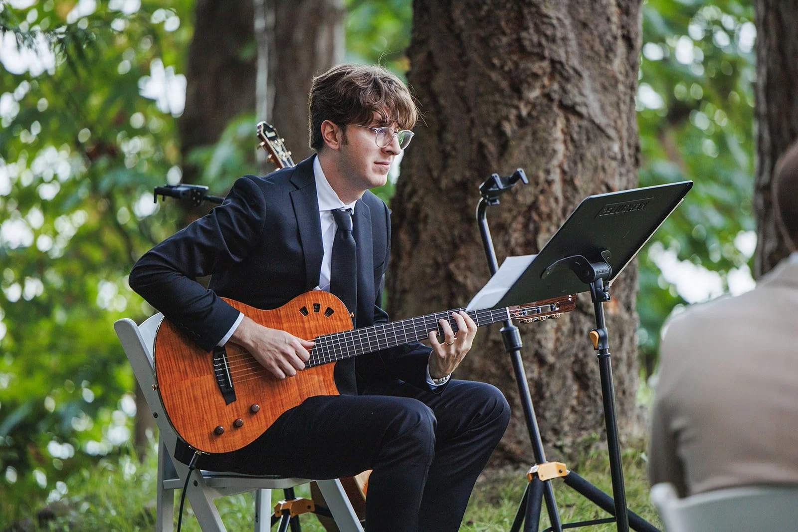 A man in a black suit and glasses playing an acoustic guitar during an outdoor event in a wooded area.