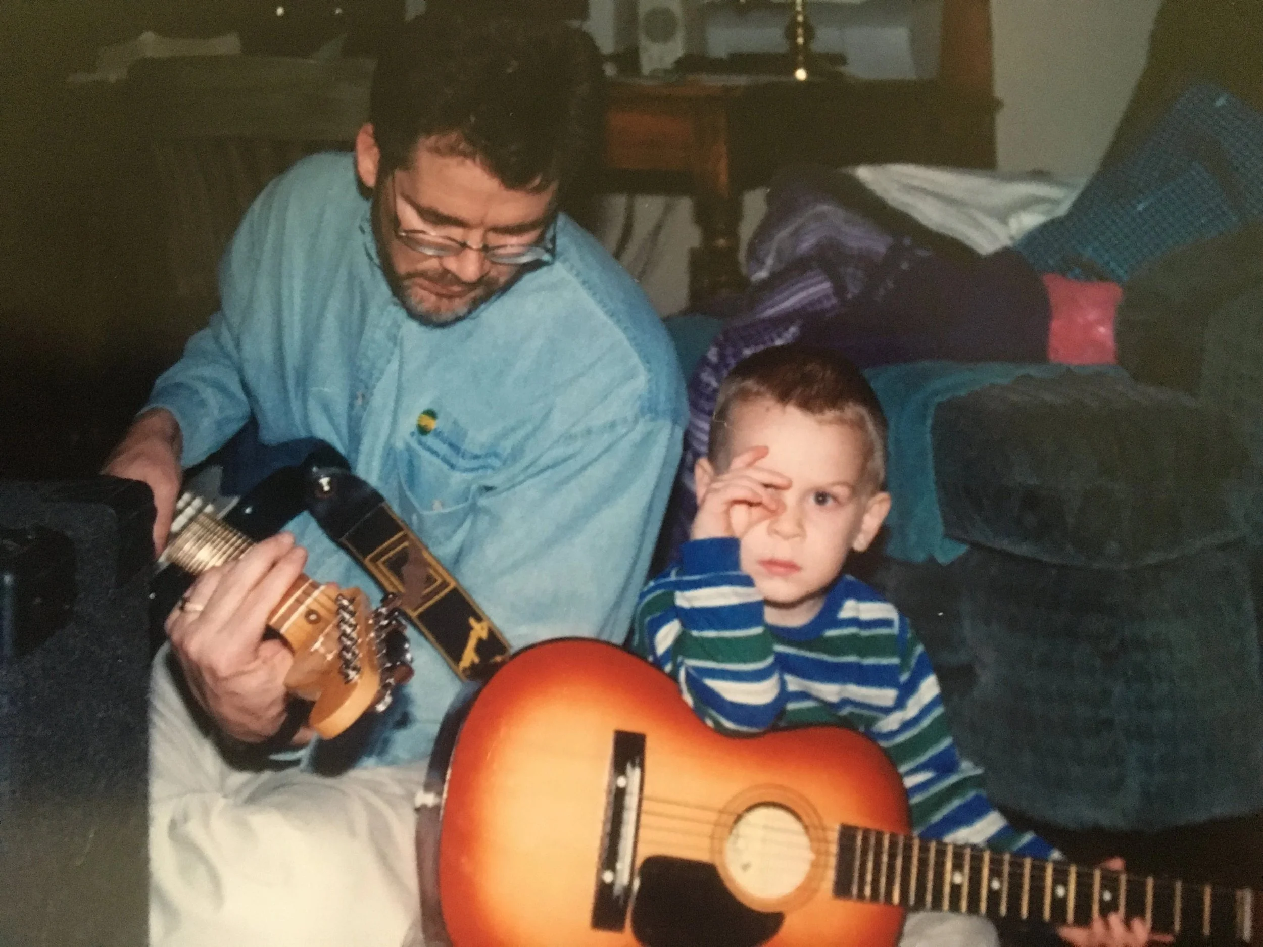 A man with glasses and a beard, wearing a light blue shirt, playing an acoustic guitar next to a young boy with short hair, wearing a striped shirt, holding a second guitar and making a playful gesture with his hand near his eye while sitting on the floor in a cluttered room.