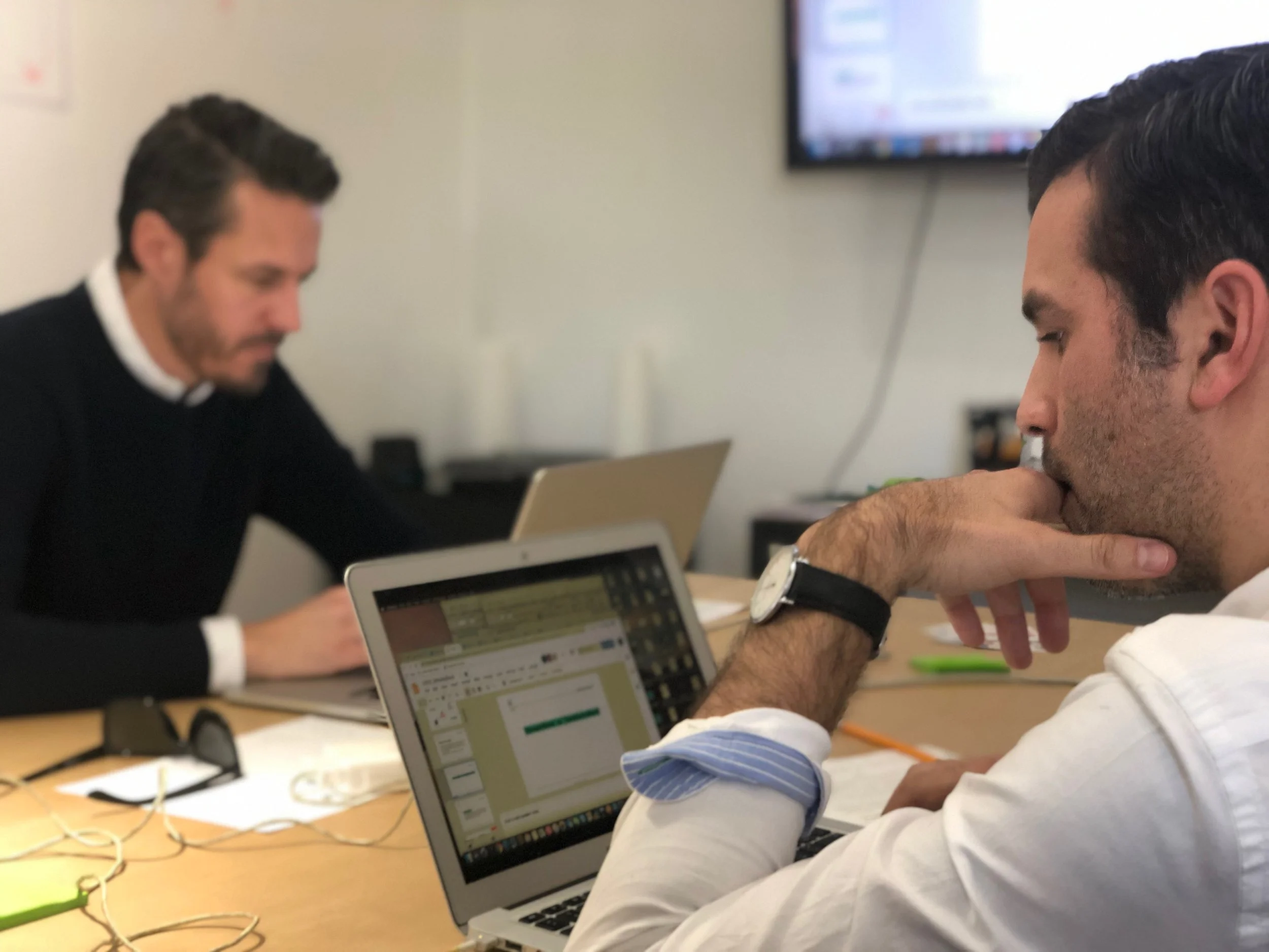 Two men sitting at a conference table with laptops and paperwork, engaged in work, in an office setting.