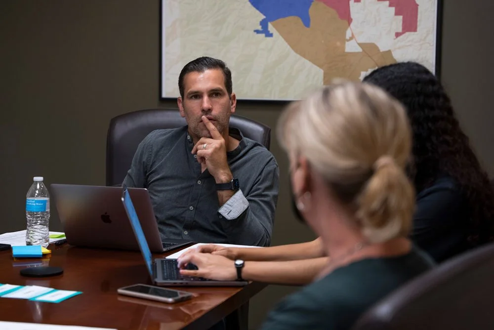 A man with dark hair and a dark gray shirt sitting at a conference table with a laptop, with his finger on his lips, thinking. Two women sit across from him, one with blonde hair in a ponytail working on a laptop, and the other with dark curly hair. There are water bottles, business cards, and electronic devices on the table, and a map or diagram on the wall behind them.