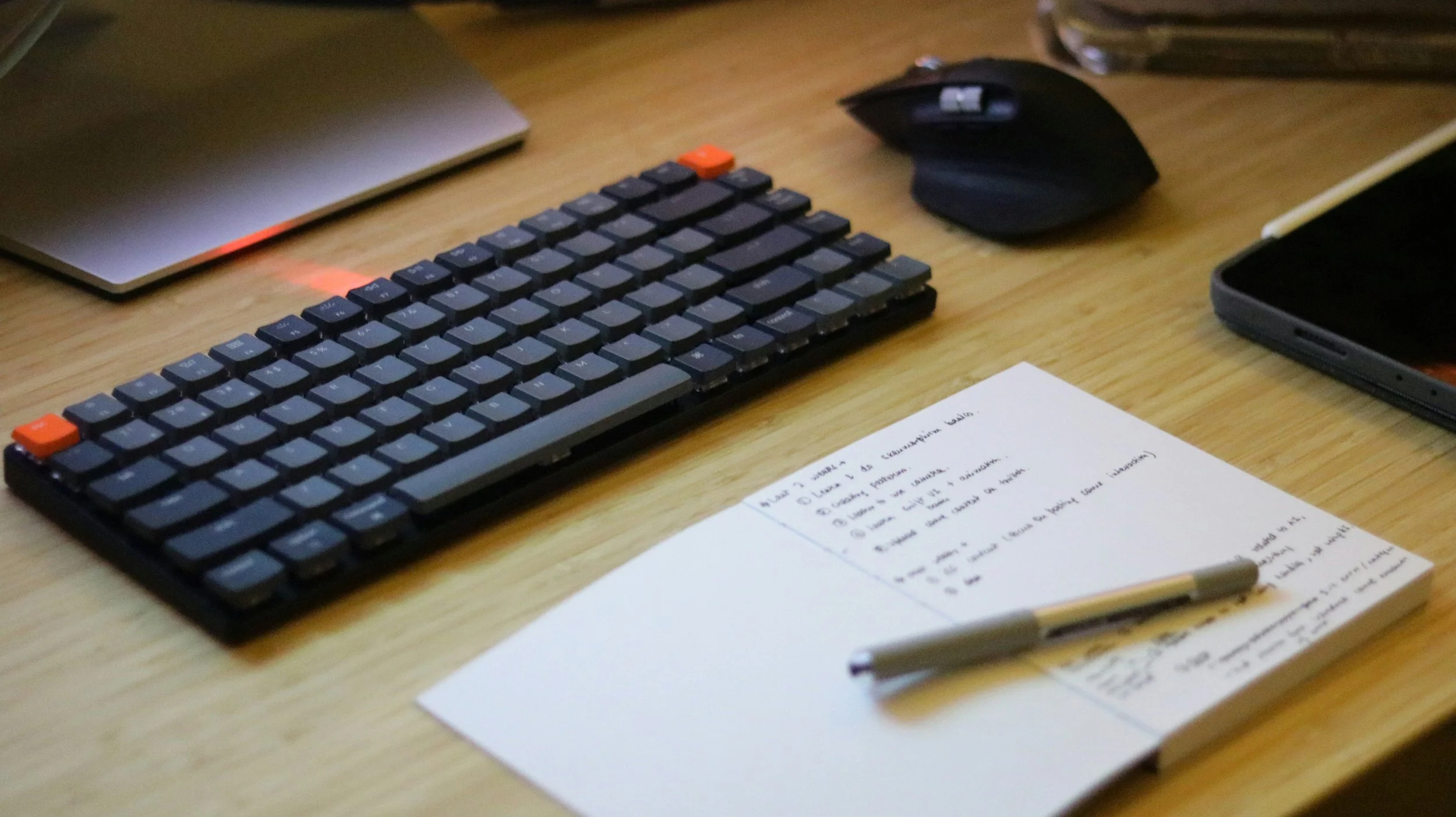 A computer keyboard, mouse, smartphone, notepad with handwritten notes, and a pen on a wooden desk.