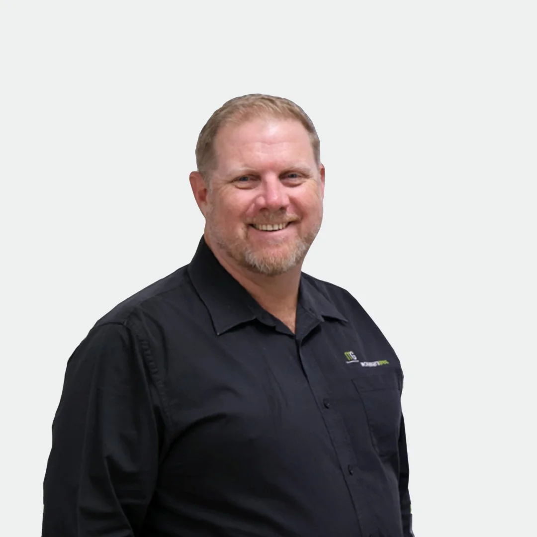 A smiling man with short, light-colored hair and a beard, wearing a black collared shirt, standing against a plain light background.