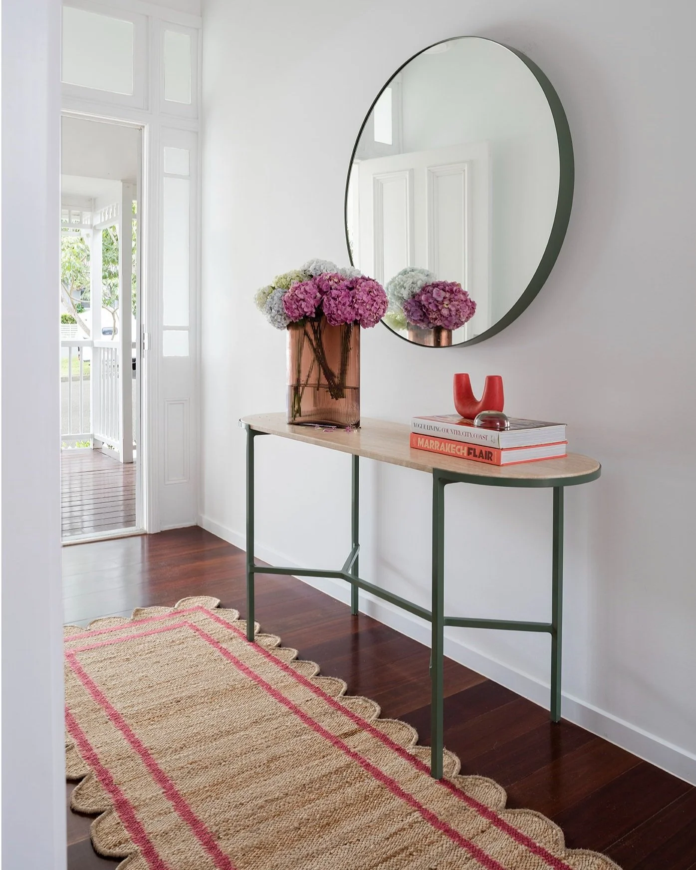 The entry of an architect designed queenslander renovation featuring a fun scalloped rug with red piping, and green metal entry tale and matching round mirror. Colourful accessories, large rust vase and blooms of purple and green hydrangeas