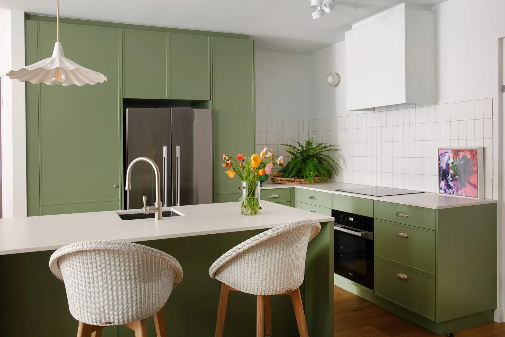 A kitchen in Green - Dulux Pesto cabinetry, white wicker bar stools, a ceramic fluted white light and white benchtop and white tile splashback,