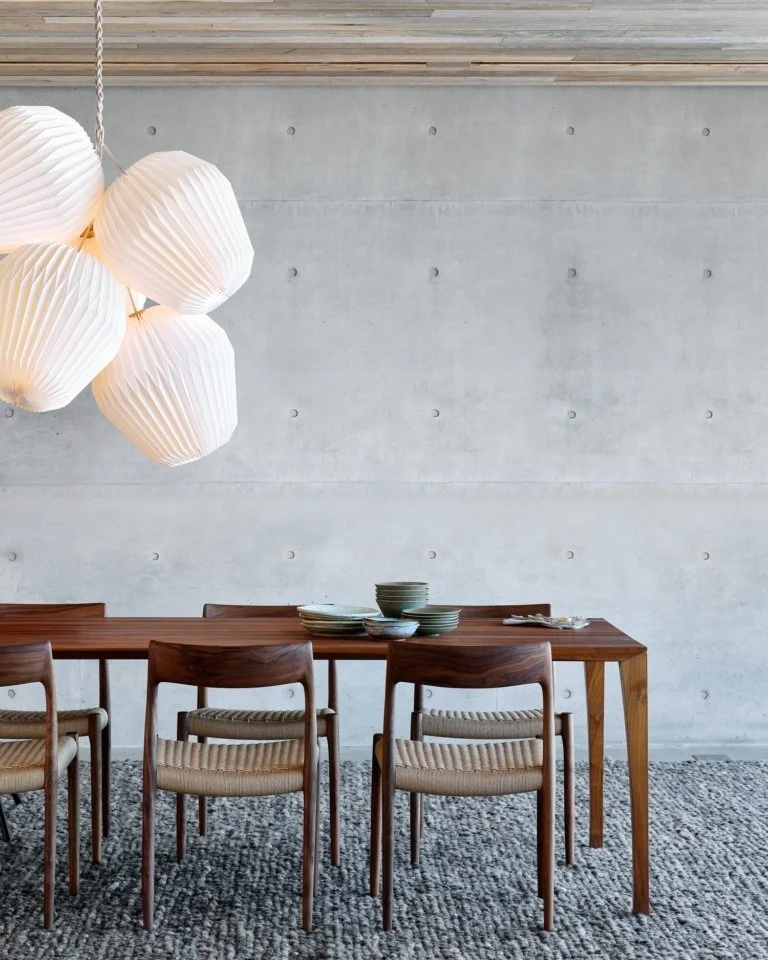 large paper pleated pendant light from Great Dane Furniture hangs over a Danish timber dining table and dining chairs with a grey wool rug and concrete wall