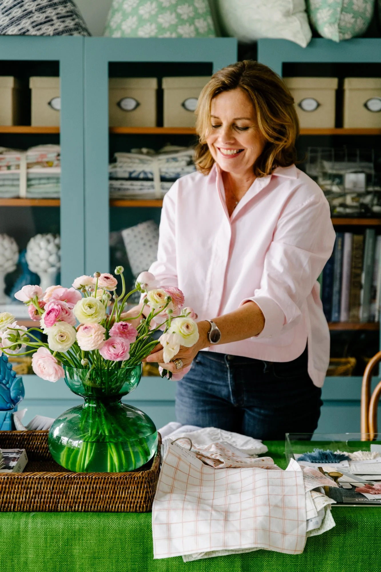 A woman arranging a bouquet of pink and white ranunculus flowers in a green glass vase on a green table.