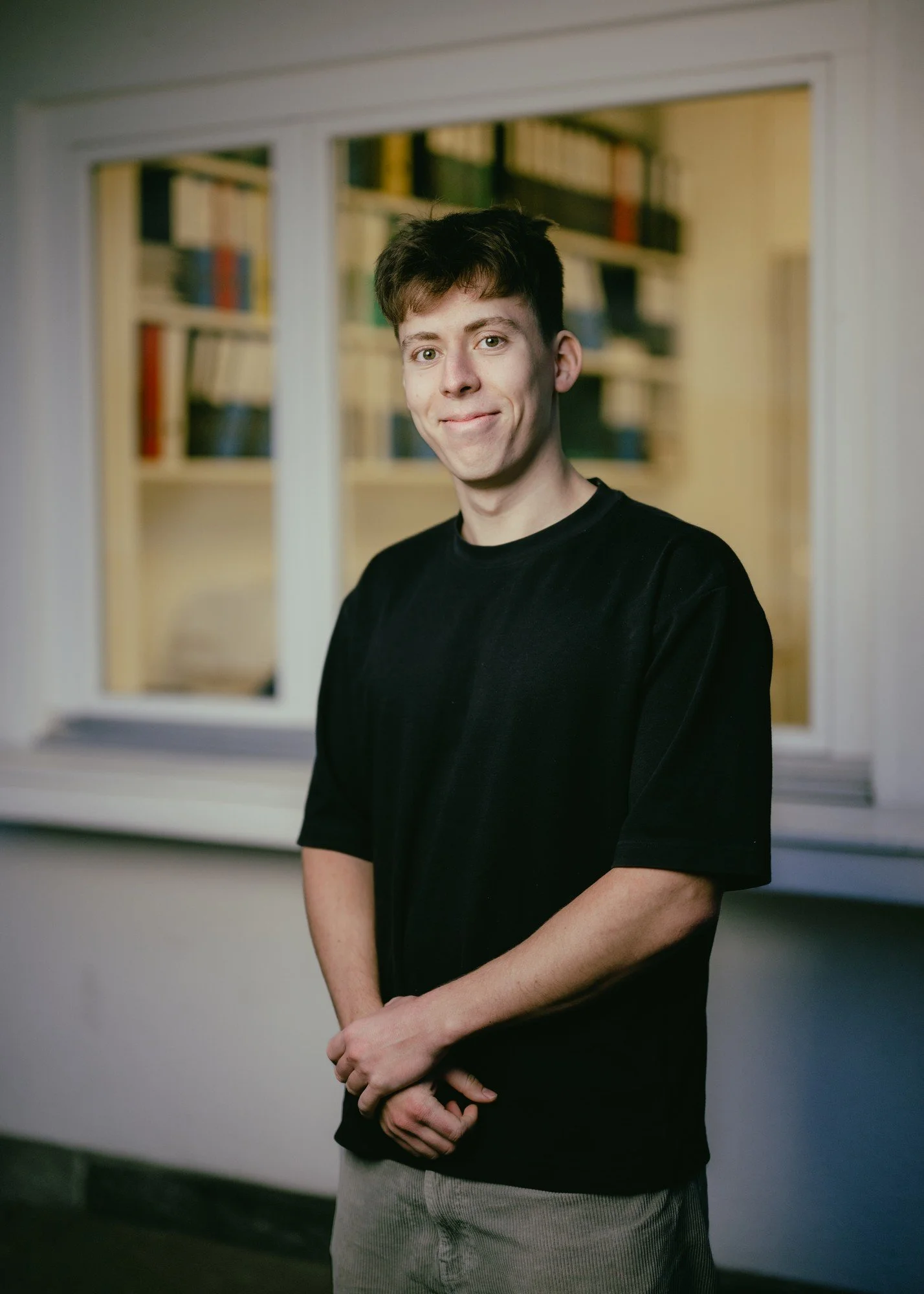 Ein junger Mann mit schwarzem T-Shirt der vor einem Fenster steht und lächelt.