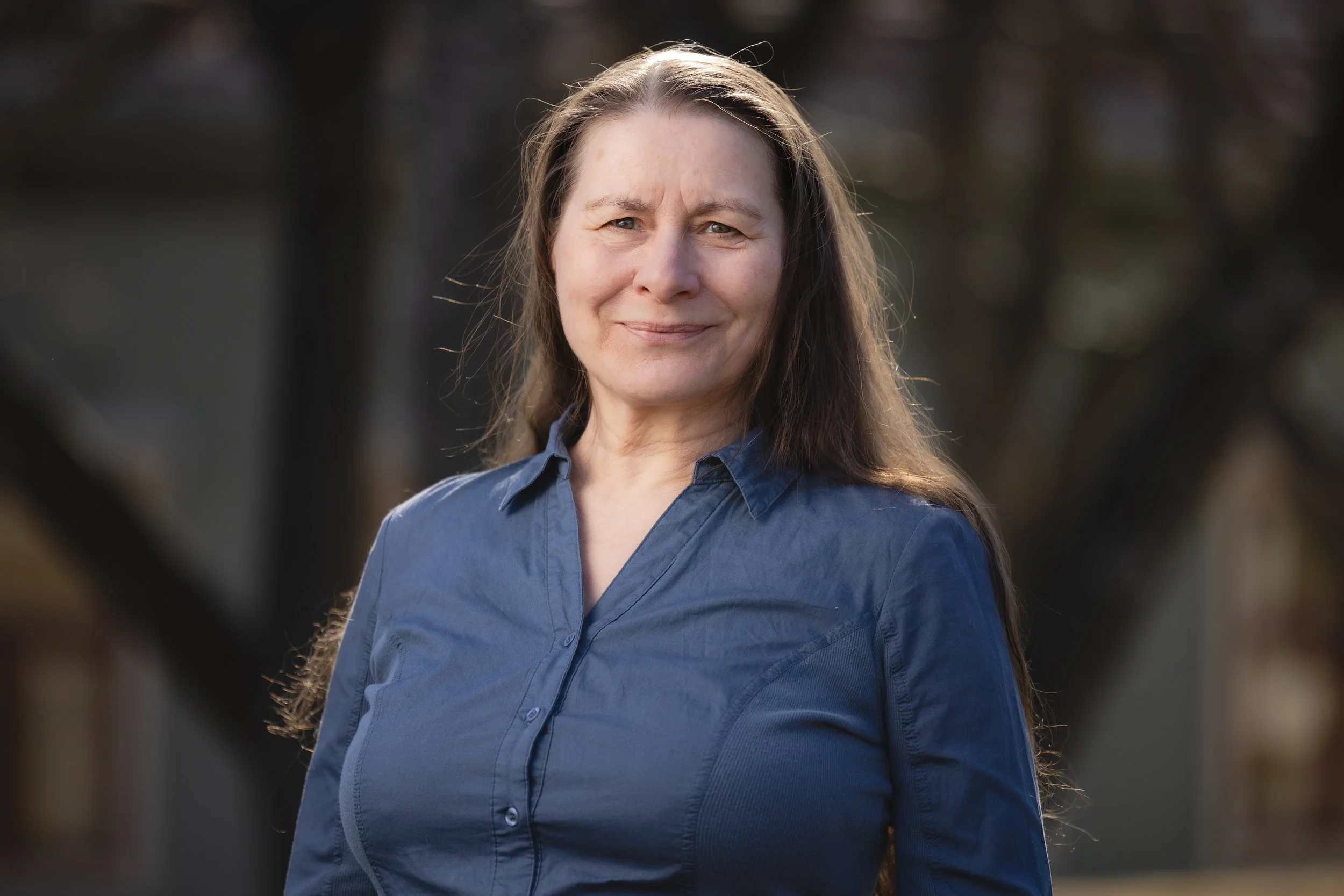A woman wearing a blue button-up shirt standing outdoors with a blurred background of trees and buildings.