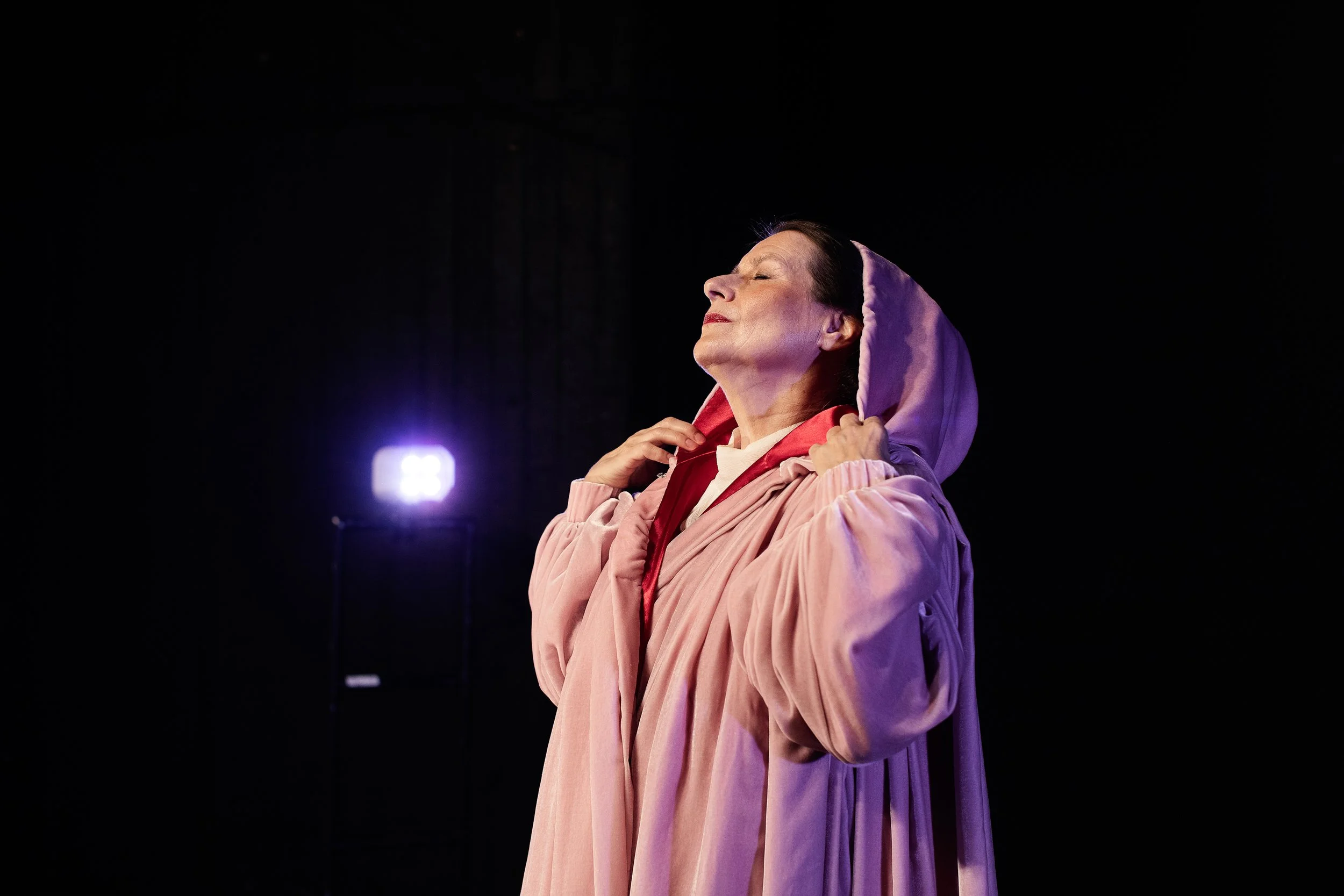 Woman with short dark hair wearing a pink hoodie and beige cloak, eyes closed, face tilted upwards, holding the hood of her cloak with both hands, standing on a dark stage with a purple light in the background.