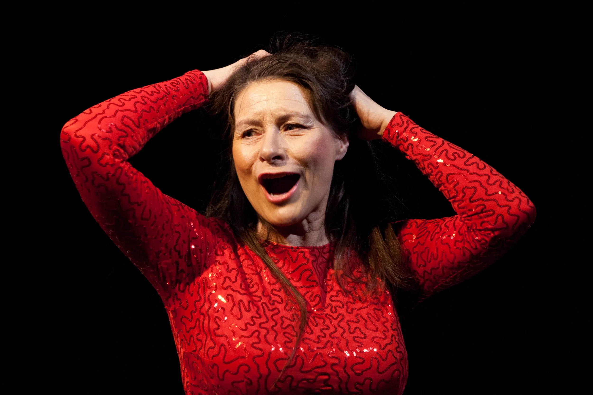 A woman with dark hair wearing a red, long-sleeved, glittery top, holding her head with her hands and appearing distressed or upset against a black background.