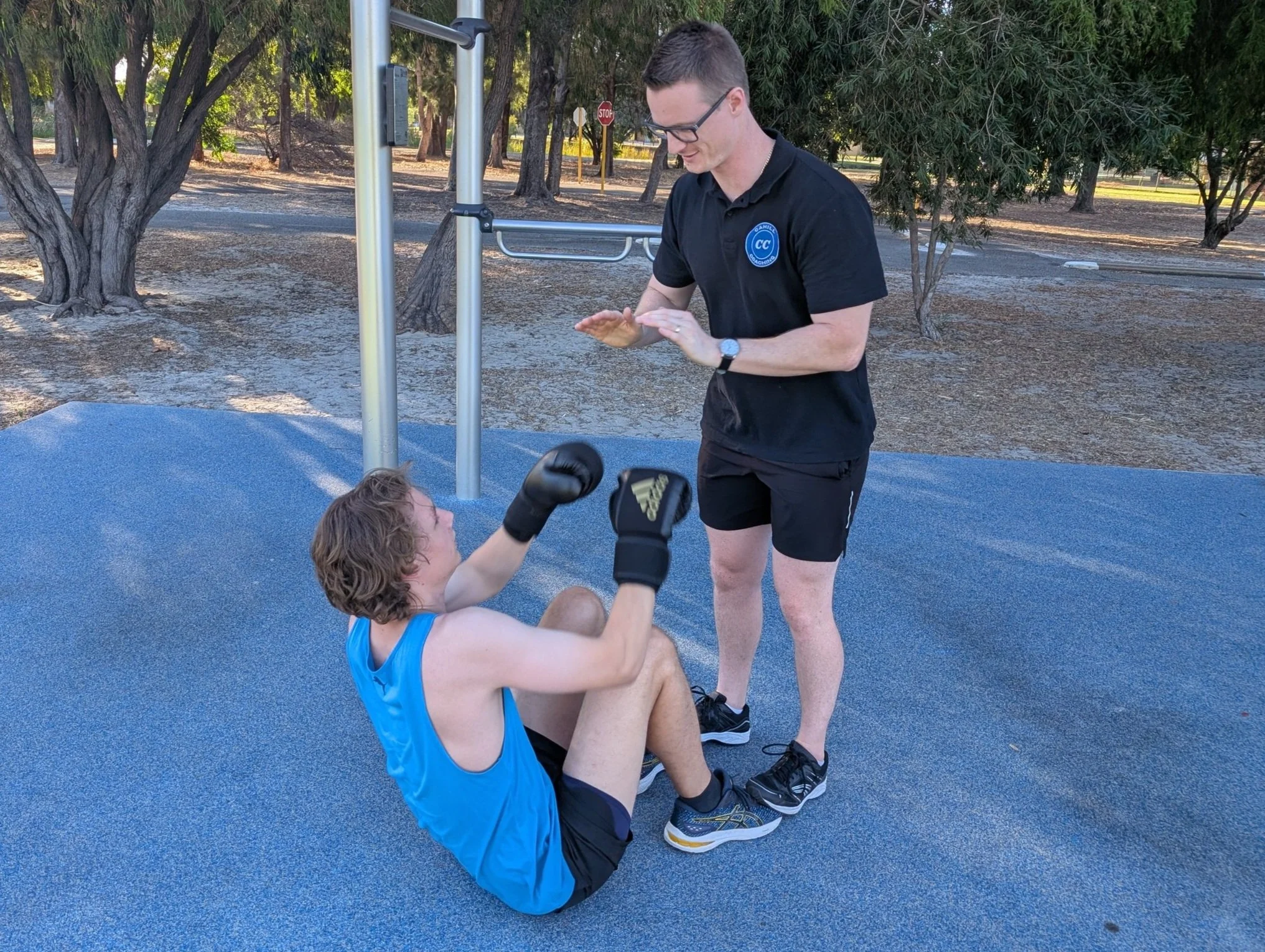 A person in a blue tank top performing sit-ups outdoors with a trainer standing nearby offering guidance. The setting is a park with trees in the background.