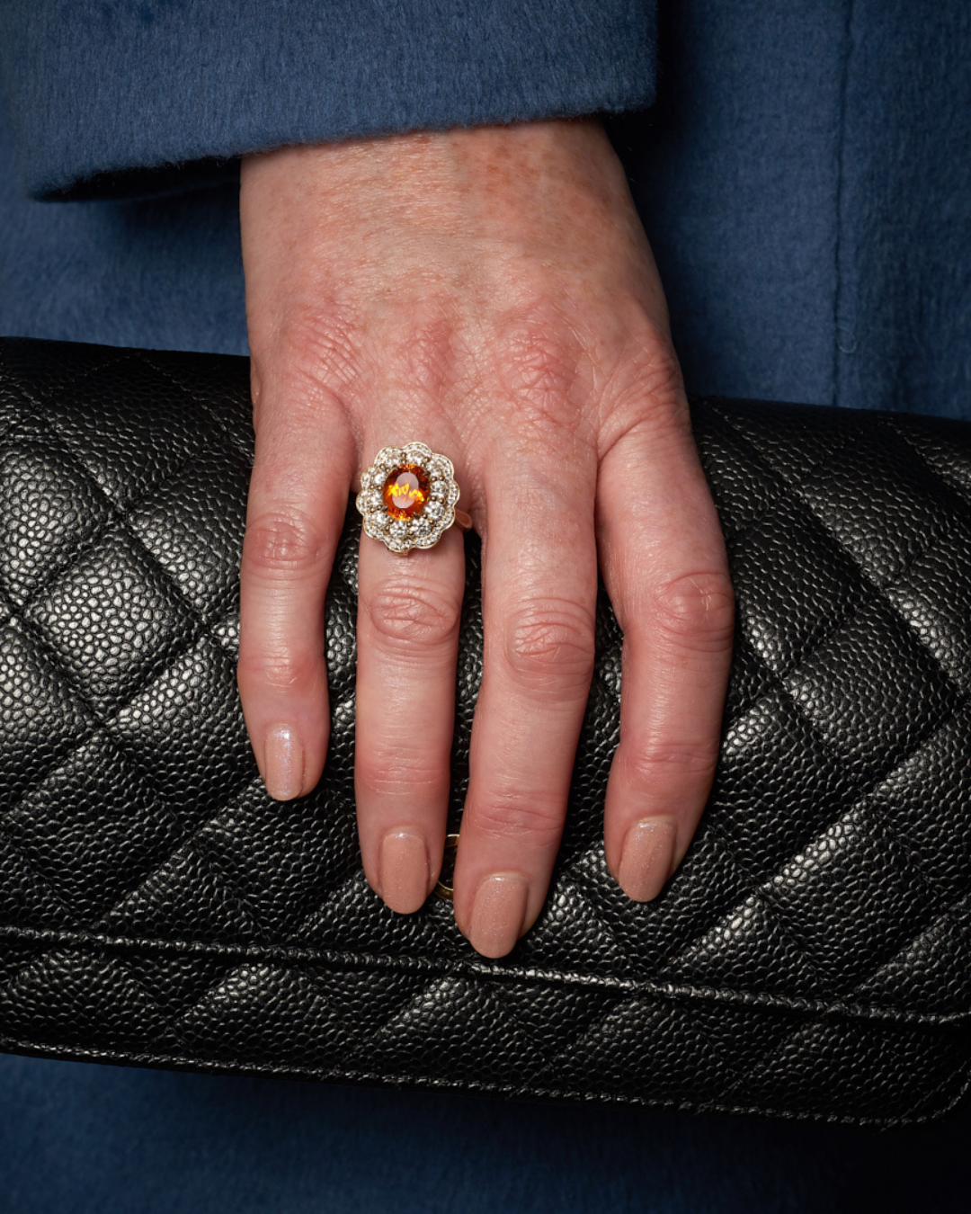 Hand resting on a black quilted purse, wearing a large floral-shaped ring with gold, diamonds, and a central orange gemstone.