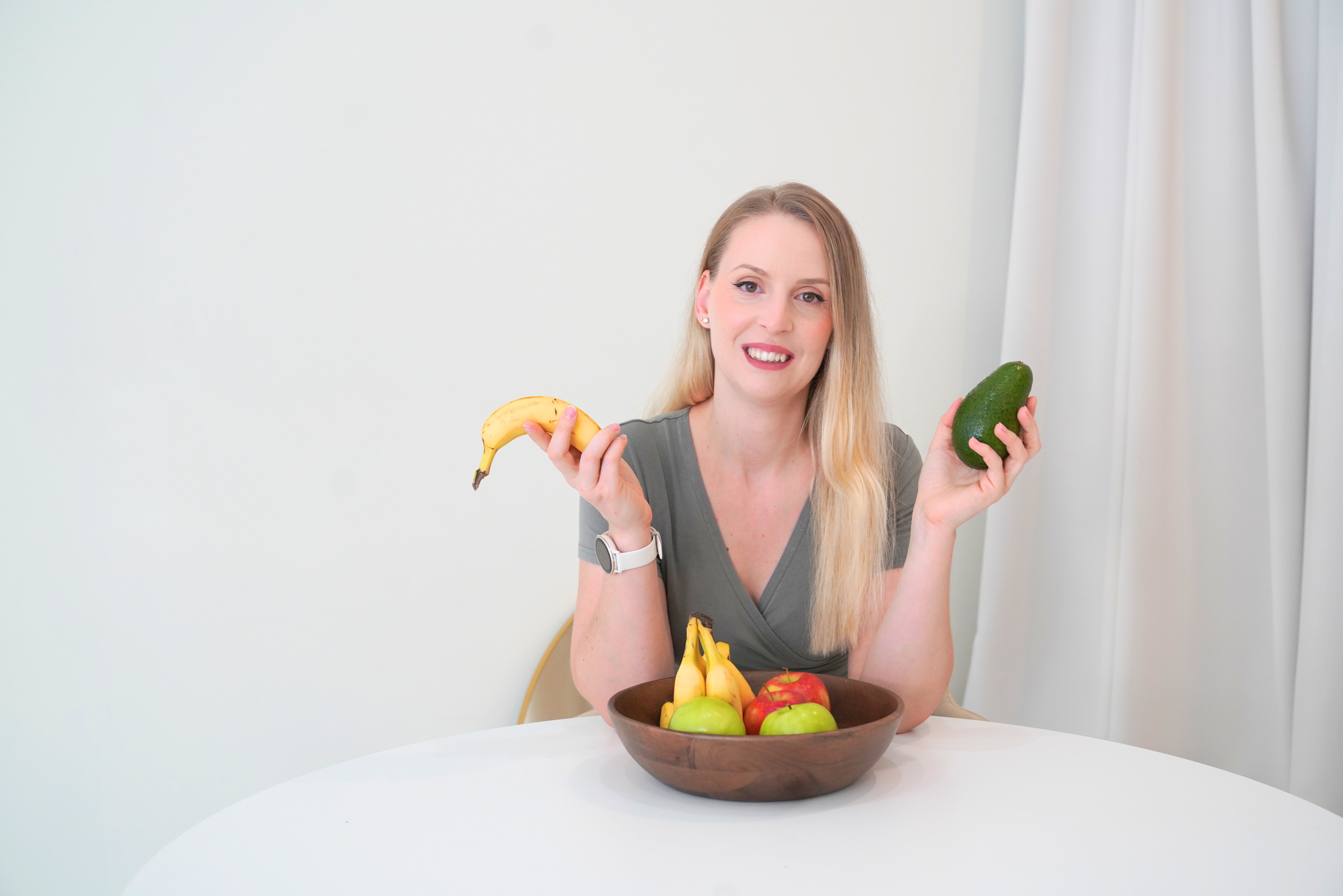 Woman with fruit sitting at a table, holding a banana and an avocado, with a bowl of assorted fruits.