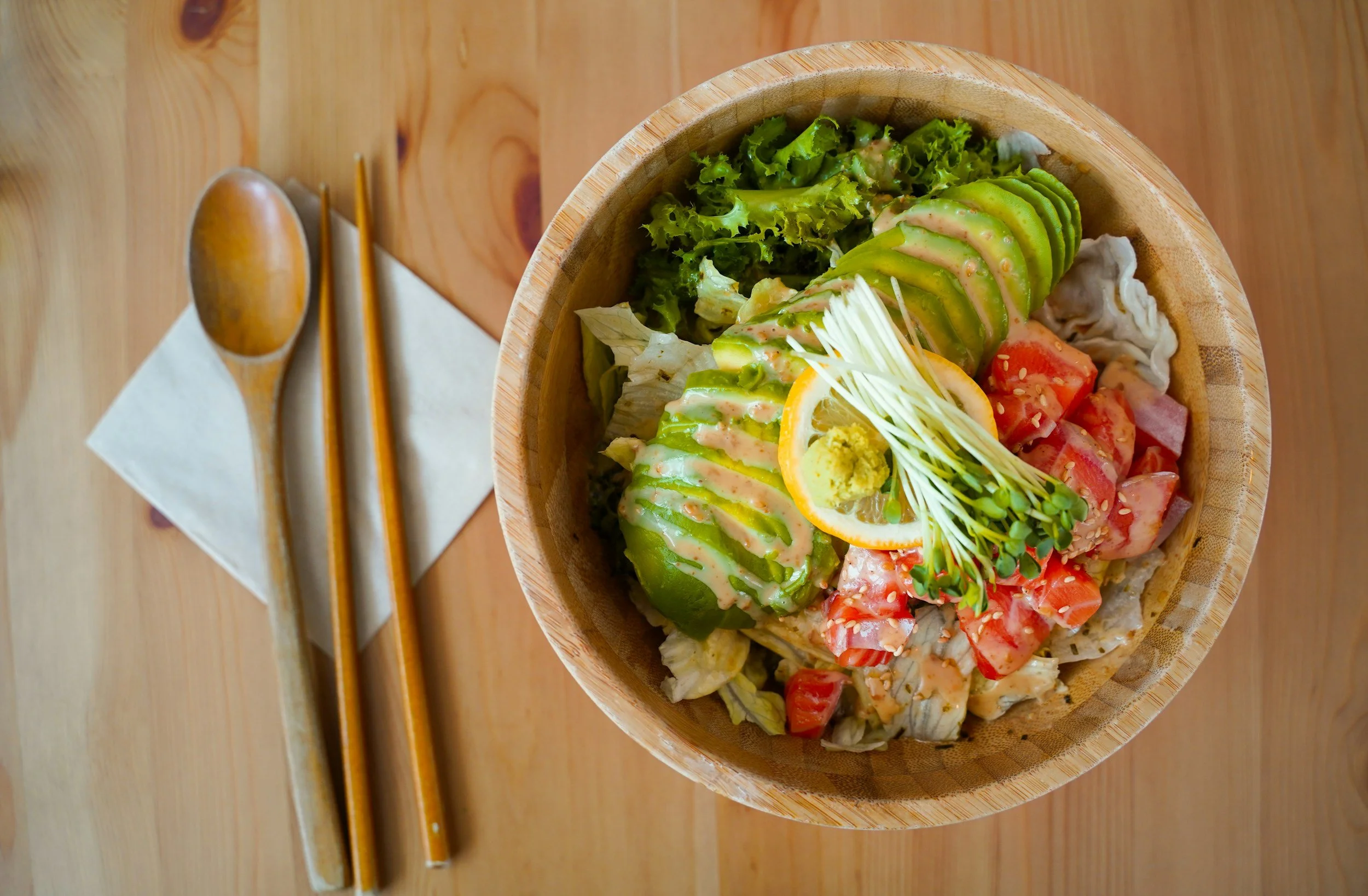 Wooden bowl of salad with sliced avocado, lettuce, tomatoes, and sprouts, garnished with a lemon slice, served with wooden spoon and chopsticks on a napkin on wooden table.