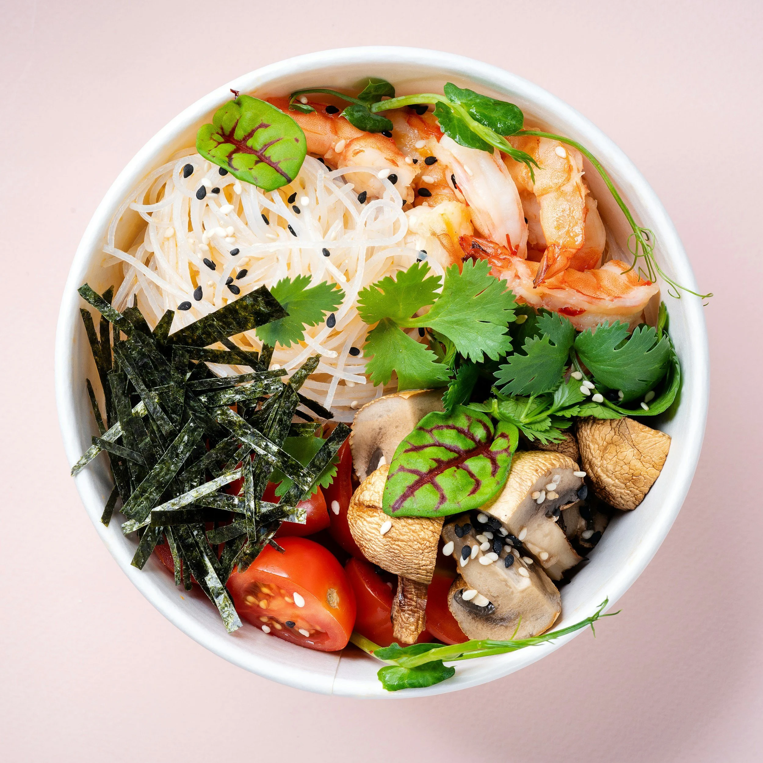 A bowl with rice noodles, shrimp, cherry tomatoes, mushrooms, seaweed strips, cilantro, and sesame seeds, garnished with leafy greens and black sesame seeds.
