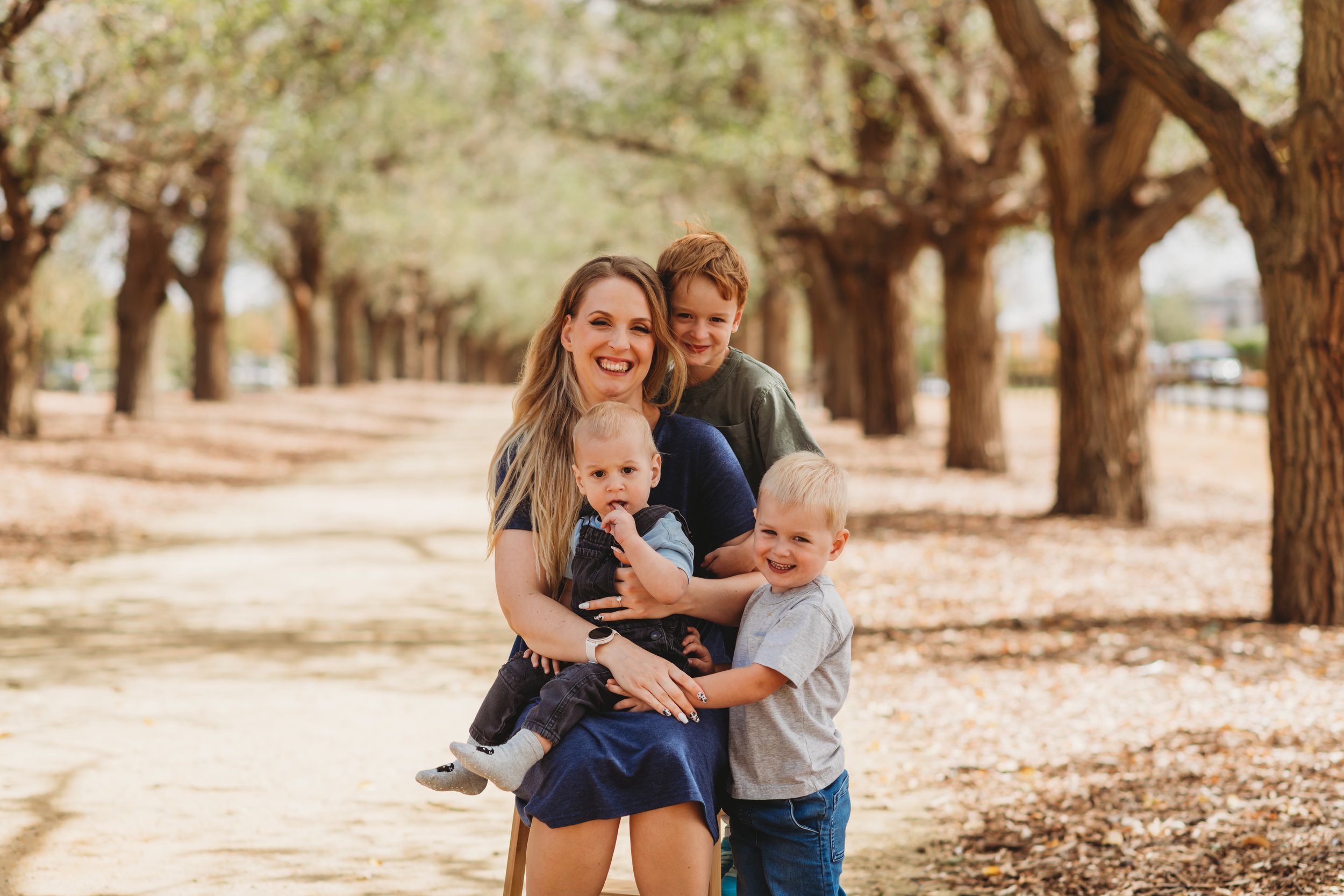 A smiling woman seated outdoors with three young children, surrounded by trees on a sunny day.