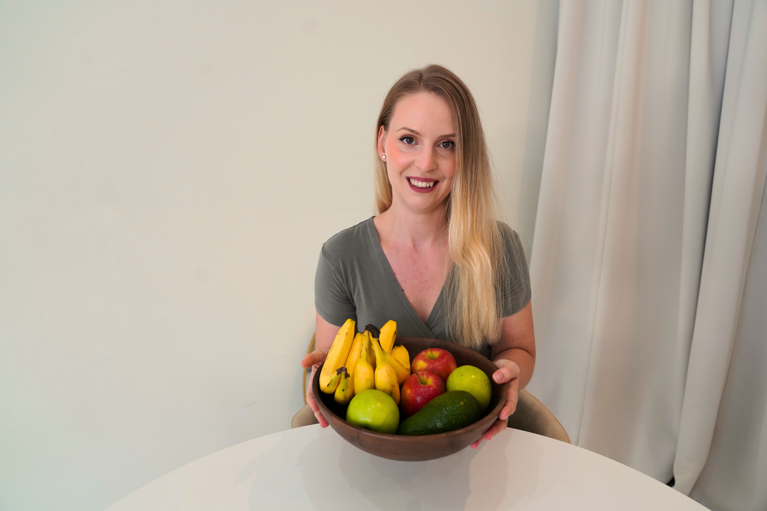 A person holding a bowl of fruit with bananas, apples, and an avocado, sitting at a table.