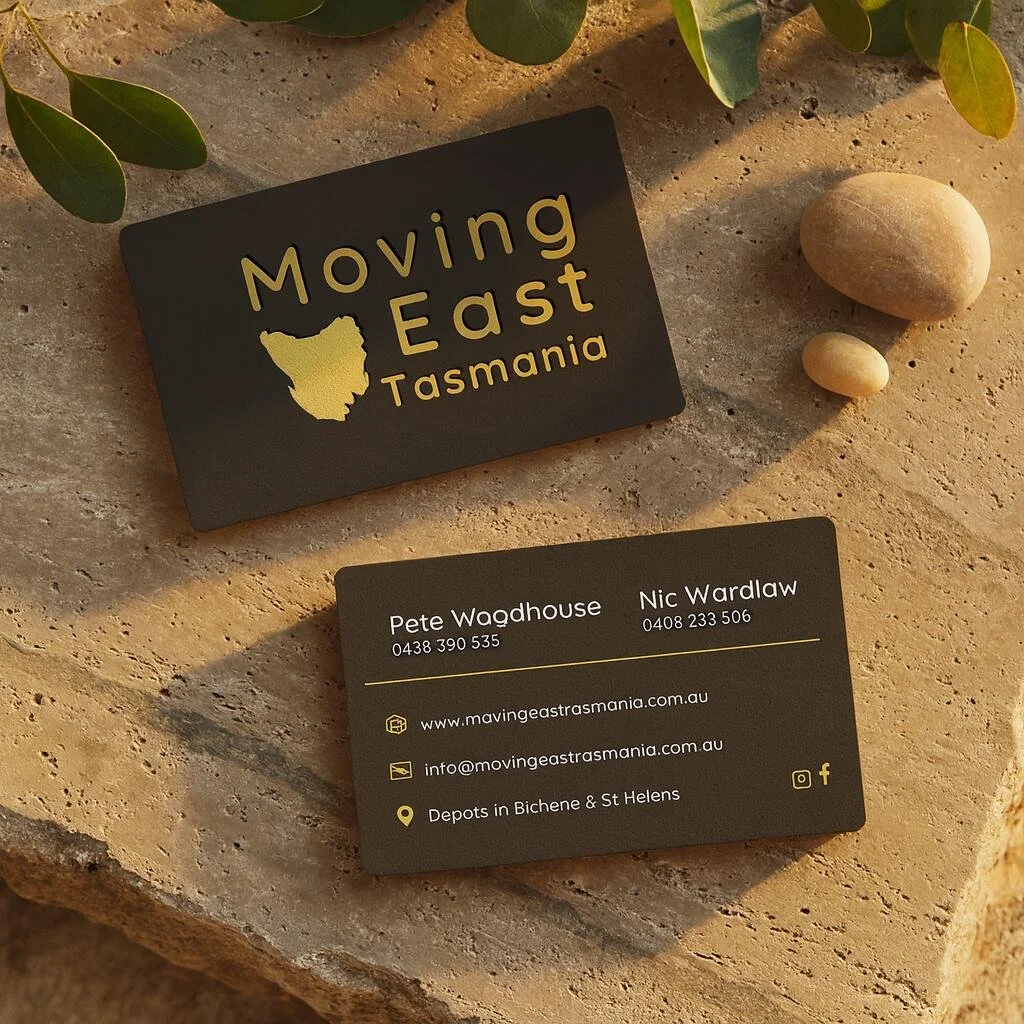 Business card for Moving East Tasmania with contact information and website, placed on a beige stone surface with two small rocks and some green foliage.