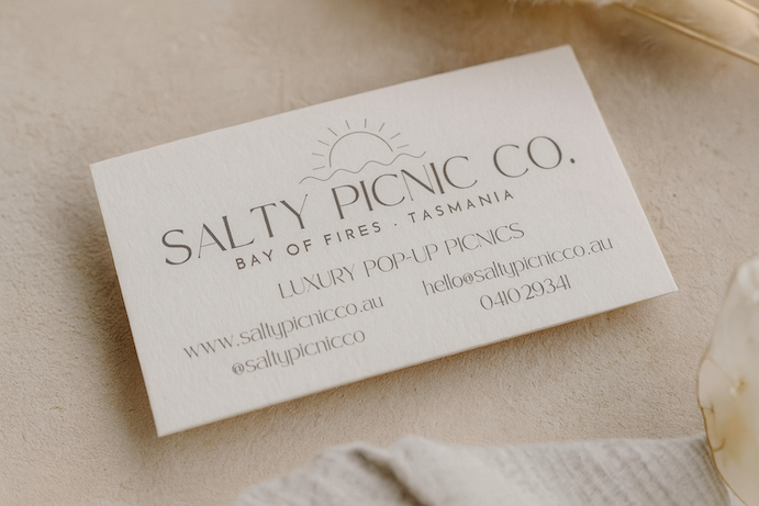 Business card for Salty Picnic Co., a luxury pop-up picnic service in Tasmania, featuring a sun and wave graphic, with contact details.
