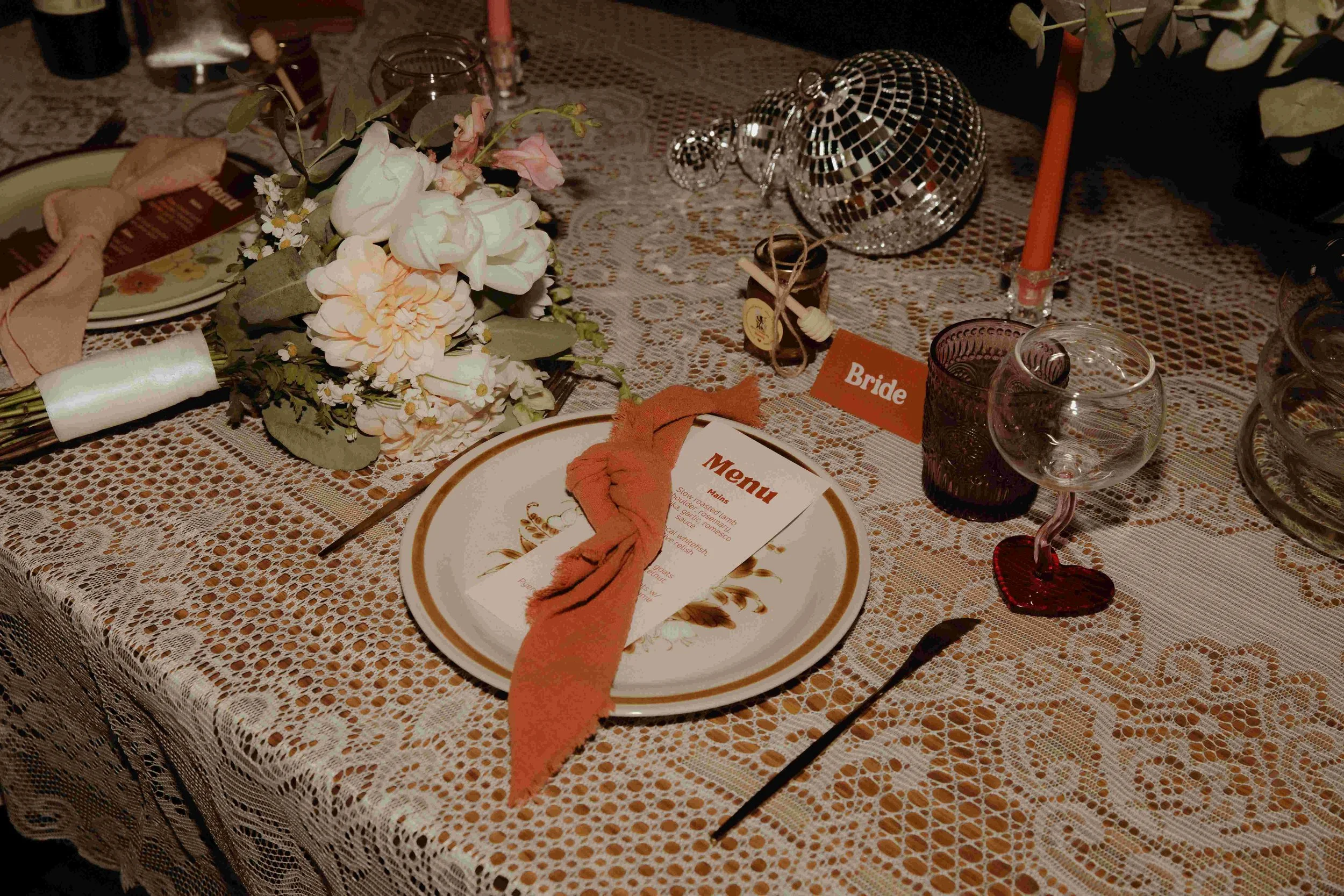 A retro themed decorated wedding reception table with a floral centerpiece, a menu card, a wine glass with a heart-shaped coaster, and a sign that says 'Bride' on a lace tablecloth.