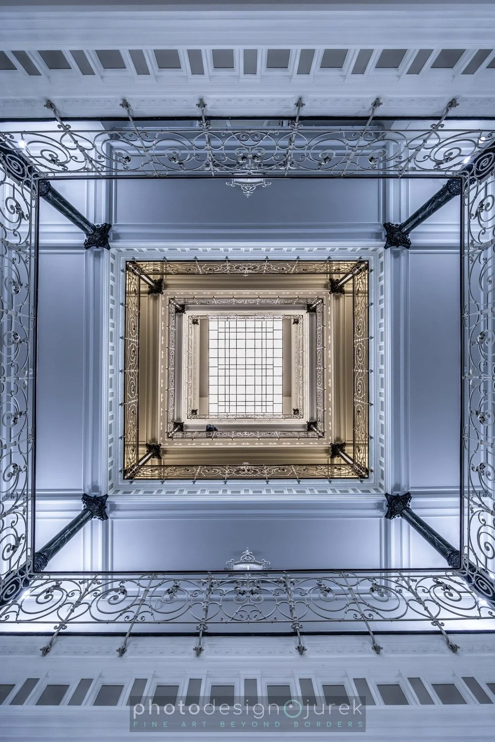 Interior view looking up a tall, square staircase with ornate white railings and black pillars, ending in a glass skylight at the top.