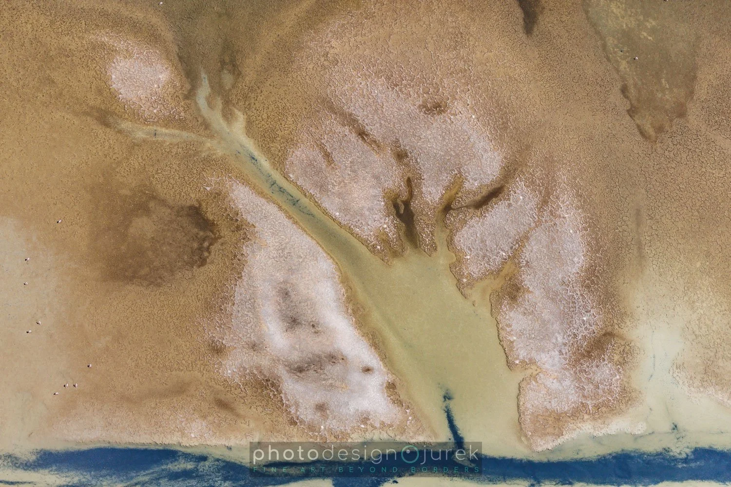 An aerial view of a desert landscape with a dry riverbed or canal running through it. The terrain includes sandy, reddish, and beige areas with patches of salt flats or dried lake beds.
