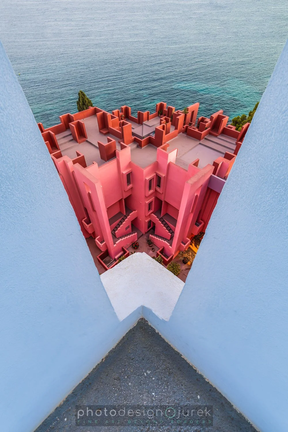 View from a high balcony looking down at a pink building with stairs on each side, overlooking a blue sea.