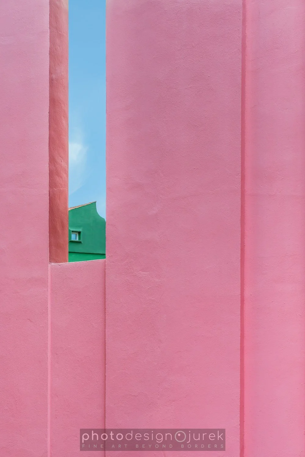 Close-up of colorful building exterior featuring large pink walls with a narrow opening showing part of a green building and a blue sky with clouds.
