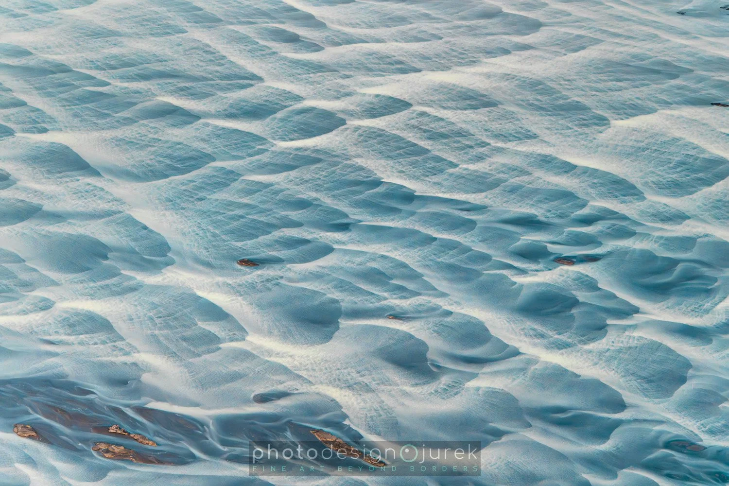 Close-up of snow-covered ground with textured and rippled surface, a few small rocks or debris scattered across the snow.