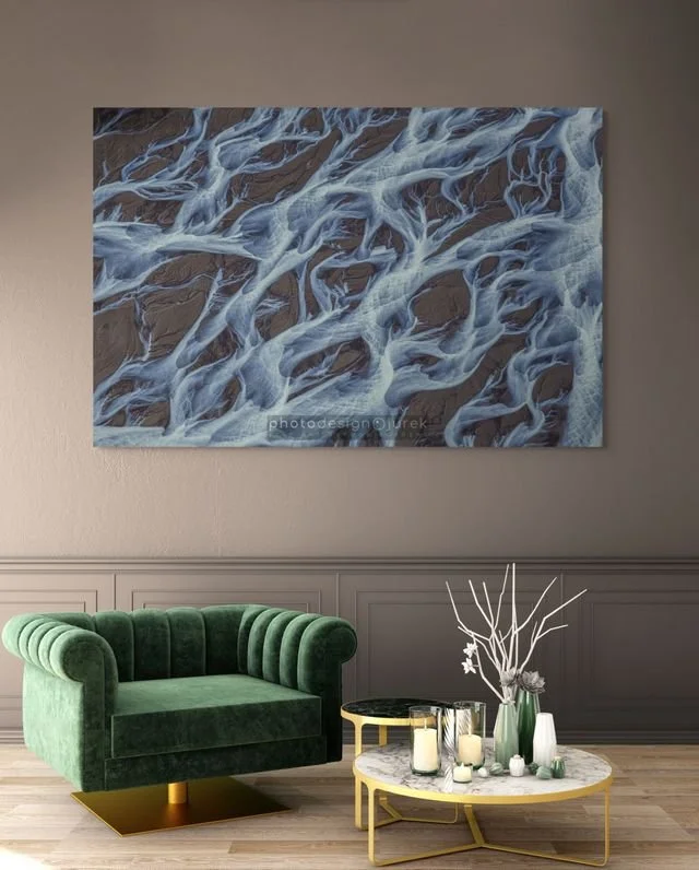 A modern interior living room with a green velvet armchair, a round marble-top coffee table, and decorative white and green vases with branches. A large abstract wall art featuring swirling blue and gray patterns hangs on a gray wall behind the furniture.