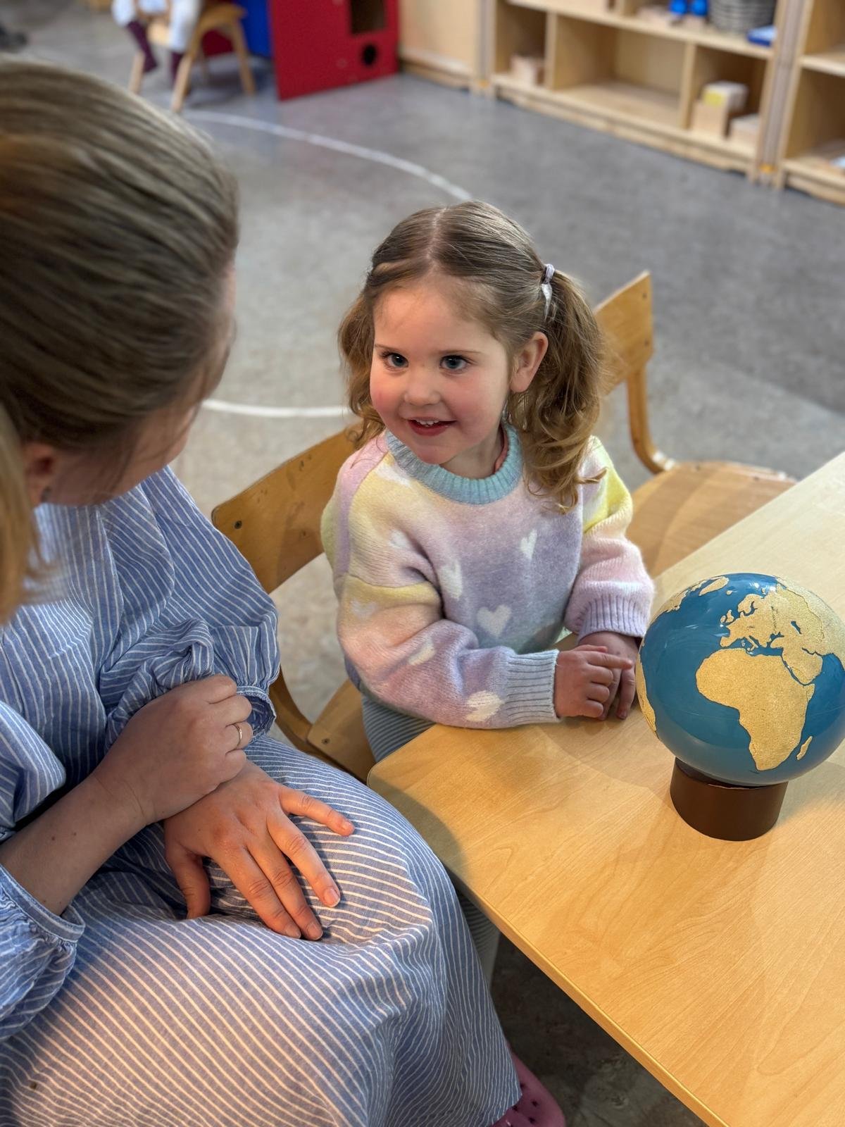 A young girl sitting at a table with an adult in a casual setting. The girl is smiling, wearing a colorful sweater and looking at a small globe on the table. The adult is in a blue-striped outfit, partially visible, showing hands on lap.
