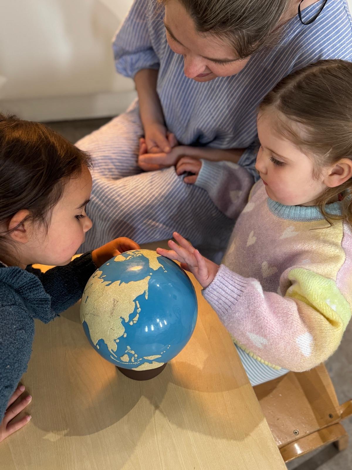 Three people, including two children and an adult, examining a globe on a table. The children are pointing and looking closely at the globe.