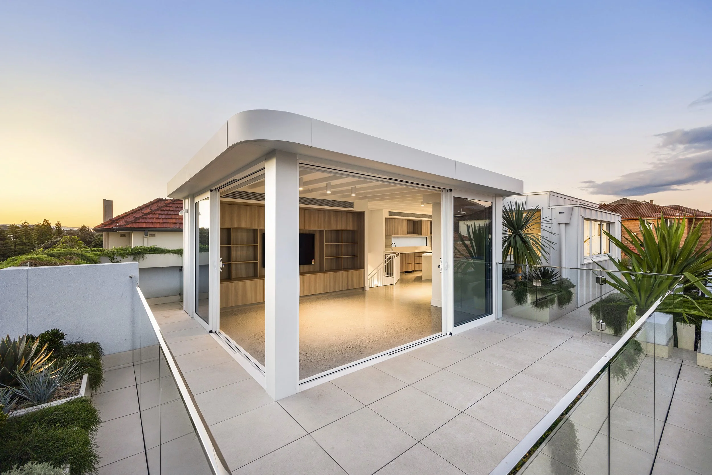 Modern rooftop balcony with glass railings, potted plants, and an open interior space with wooden shelving and kitchen area, during sunset.