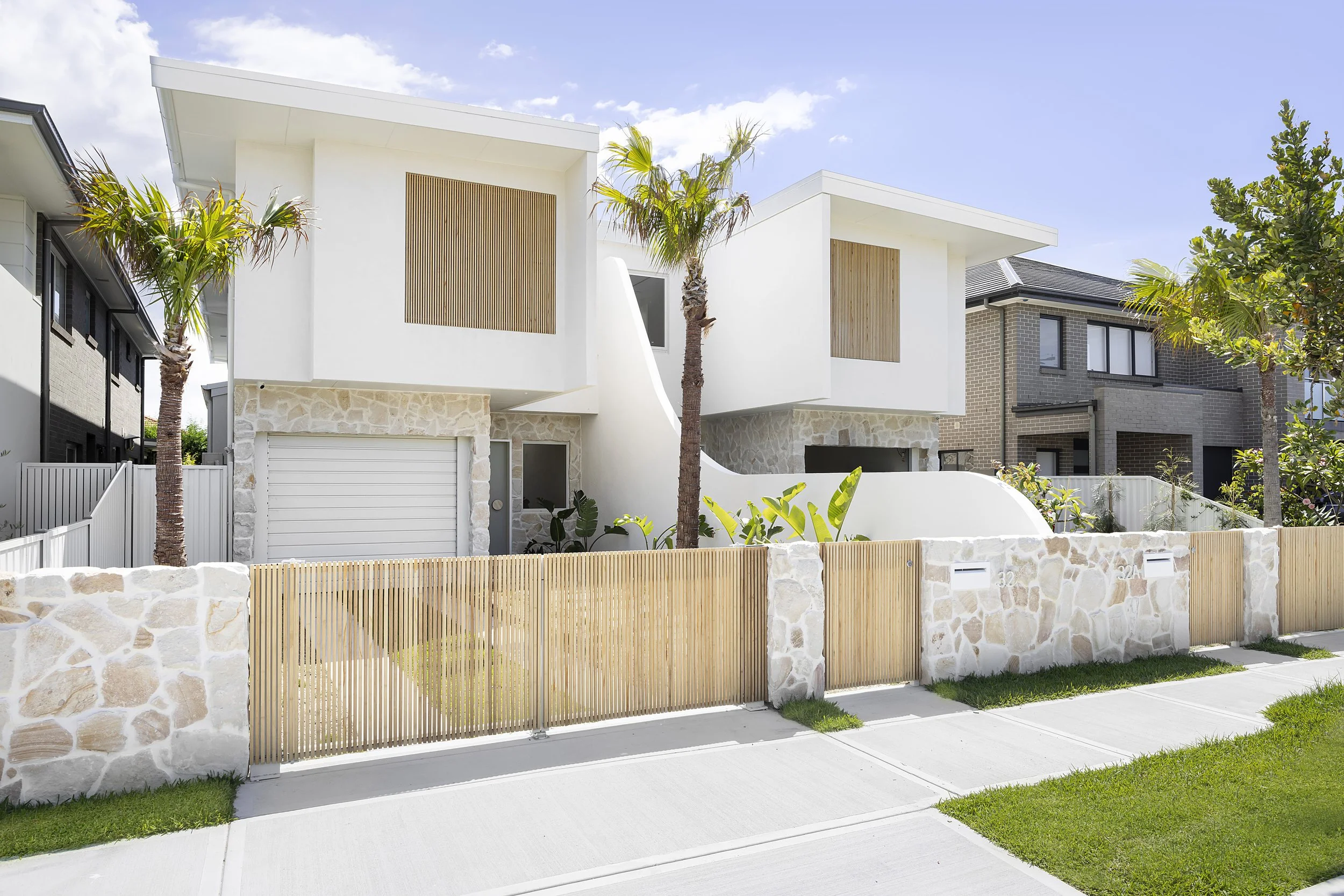 Modern duplex house with white exterior walls, stone accents, and wooden slats on upper windows. Features front driveway with white garage door, surrounded by a low stone and wooden fence, palm trees, and a green lawn.