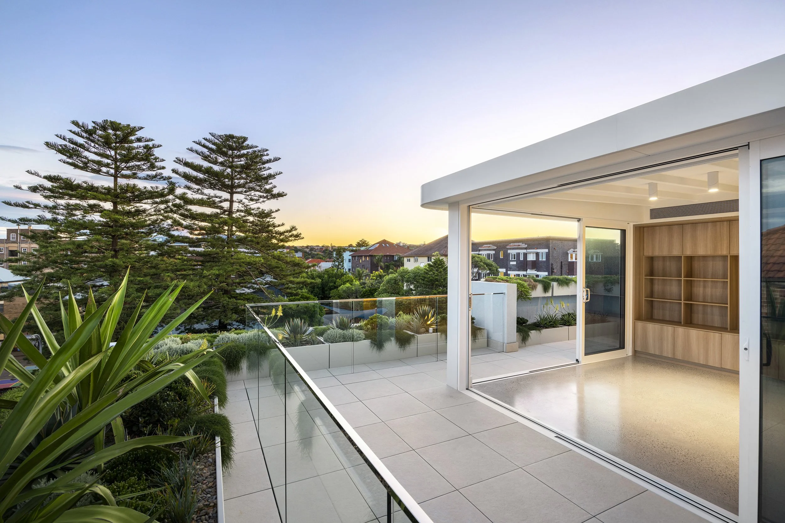 Modern balcony with glass railing, potted plants, and a view of suburban houses and trees at sunset