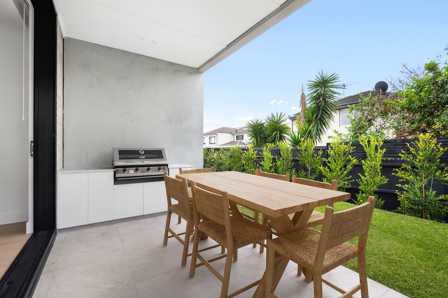 Outdoor patio with wooden dining table and chairs, built-in grill, and view of green hedge and trees under clear blue sky.