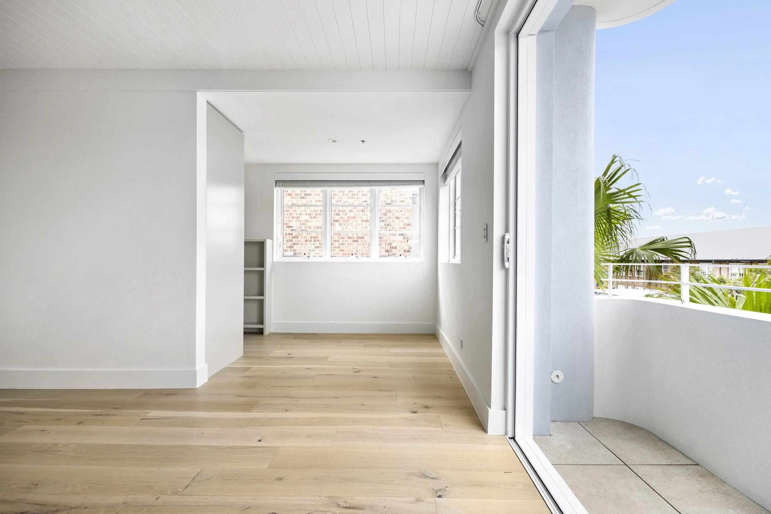 Empty room with white walls, light wood flooring, large window with blinds, and a sliding glass door leading to a balcony with a railing and green plants outside.