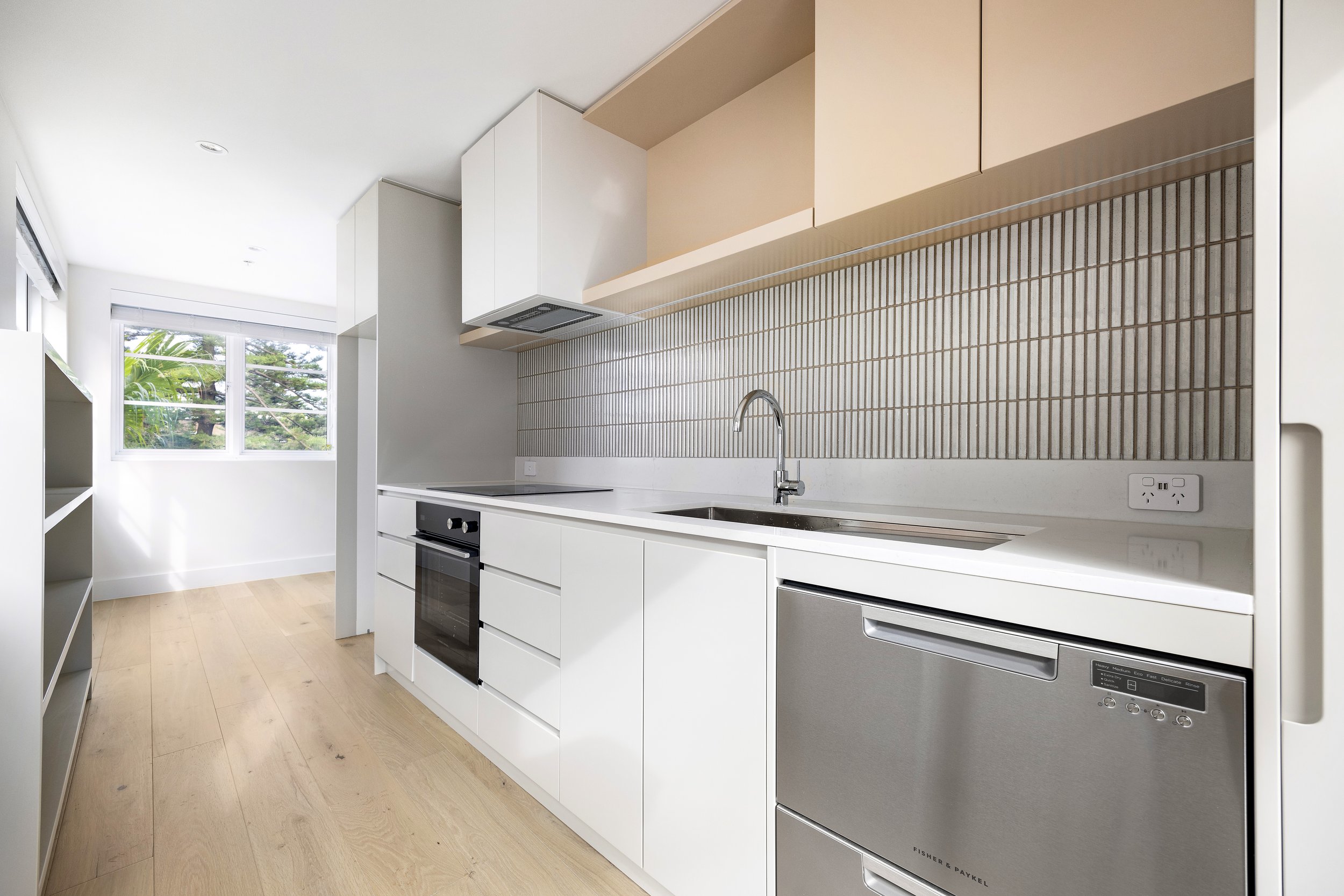 Modern kitchen with white cabinets, a black oven, a stainless steel dishwasher, a white countertop, a stainless steel sink with a faucet, and a beige tiled backsplash. A window shows greenery outside.