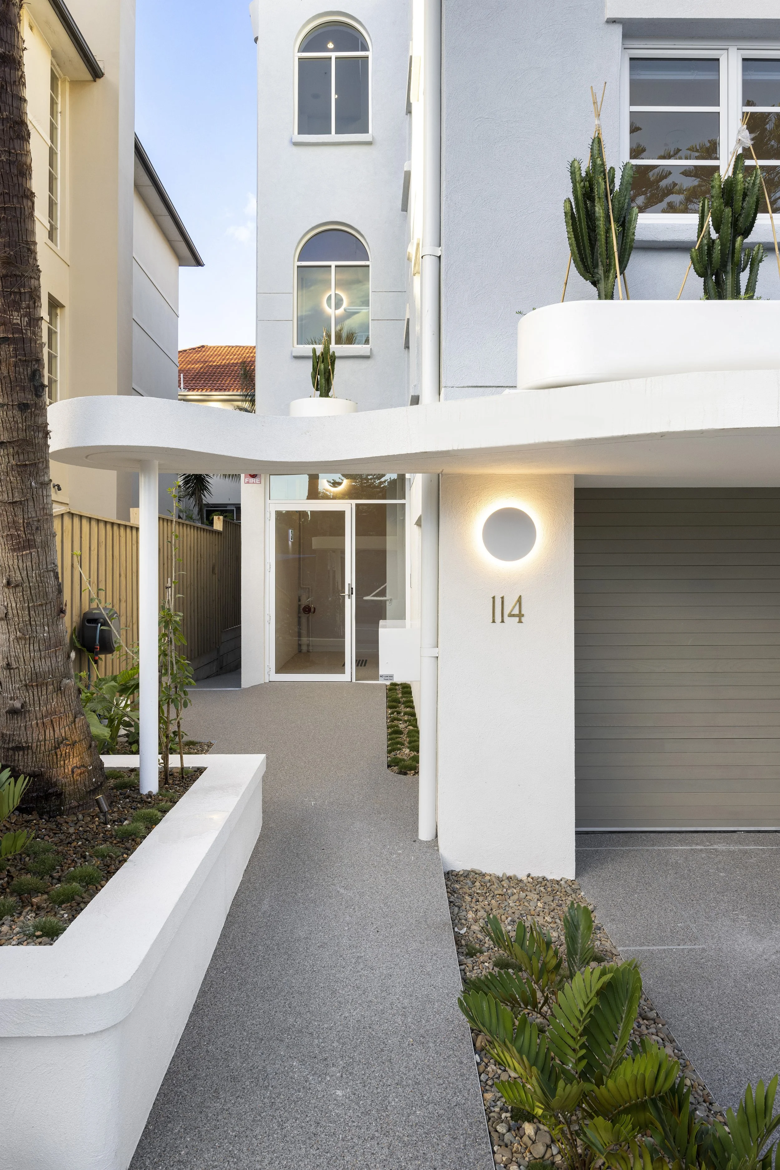 Modern white residential building with curved front entrance, featuring a walkway leading to an entrance door, decorative plants including cacti and succulents, and outdoor lighting.