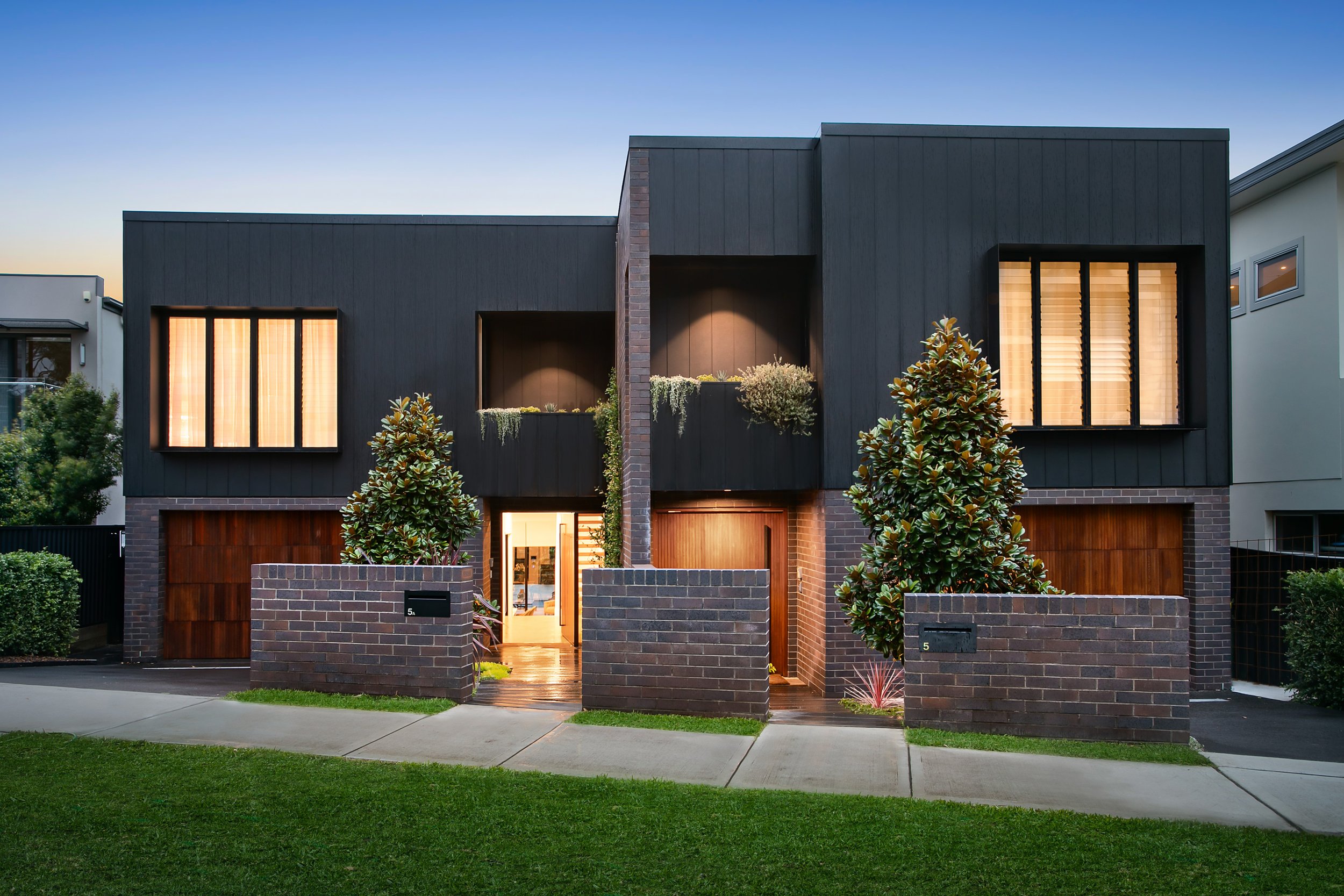 Modern two-story duplex house with dark exterior, brick accents, large windows with warm lighting, front yard with green grass, trees, and brick walls.