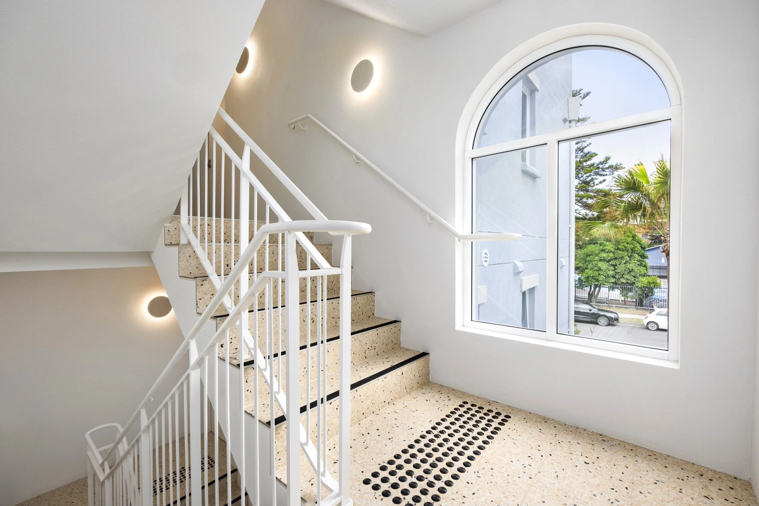 Interior stairwell with terrazzo steps, white railings, and a large arched window showing trees and parked cars outside.