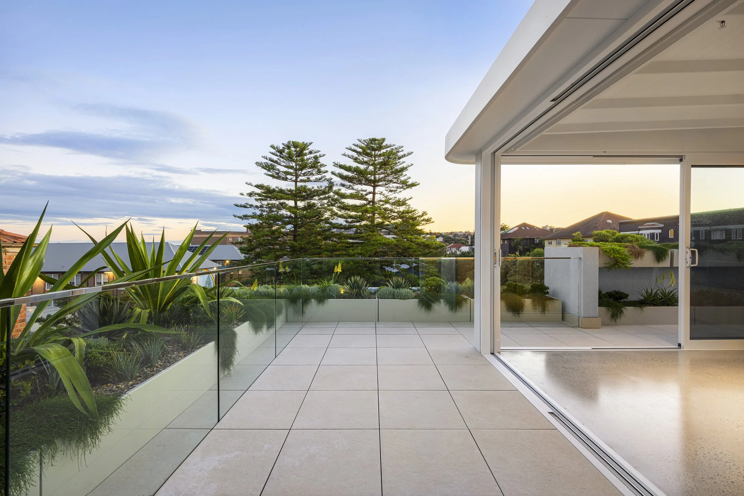 An outdoor balcony with tiled flooring, glass railing, and potted plants, overlooking a residential neighborhood with trees and houses at sunset.
