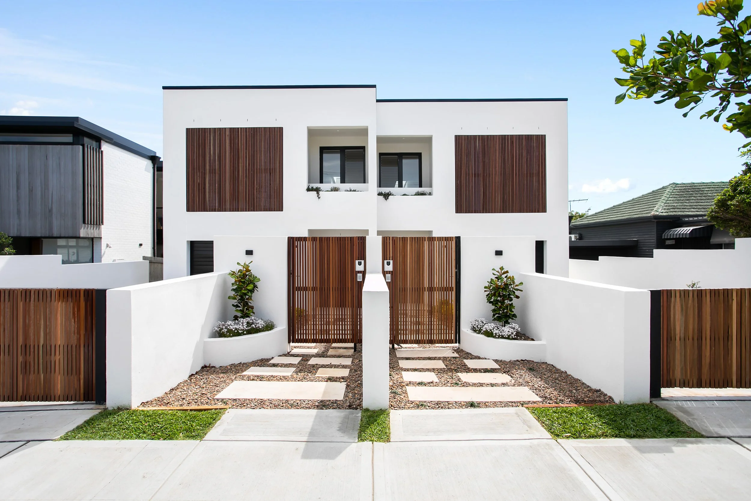 Modern white residential duplex house with timber slats, a pivot door, a central gate, and landscaped front yard.