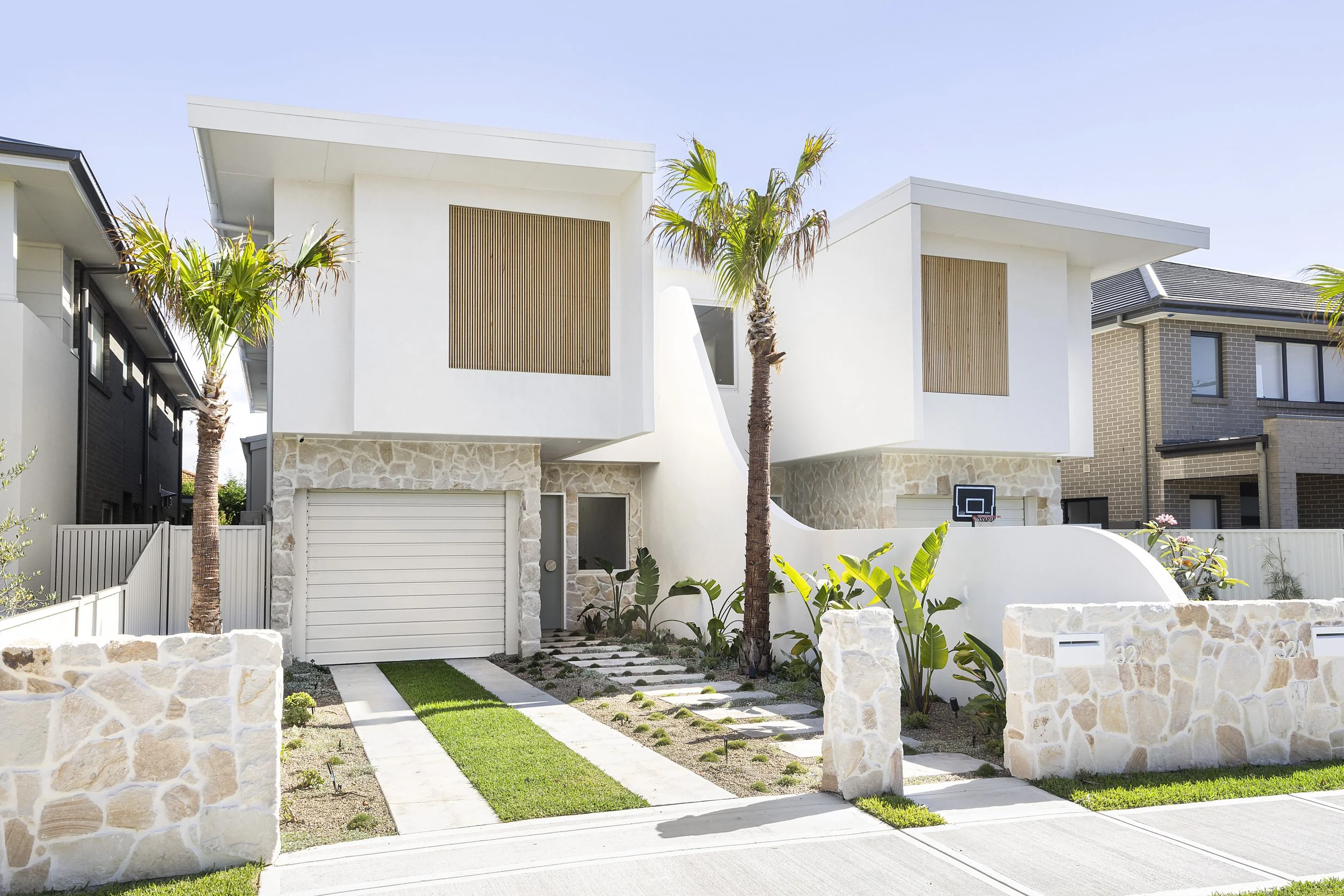 Modern two-story house with white exterior walls, stone accents on the lower level, vertical wooden slats on the upper windows, and a small front yard with palm trees, green grass strip, and a stone pathway.