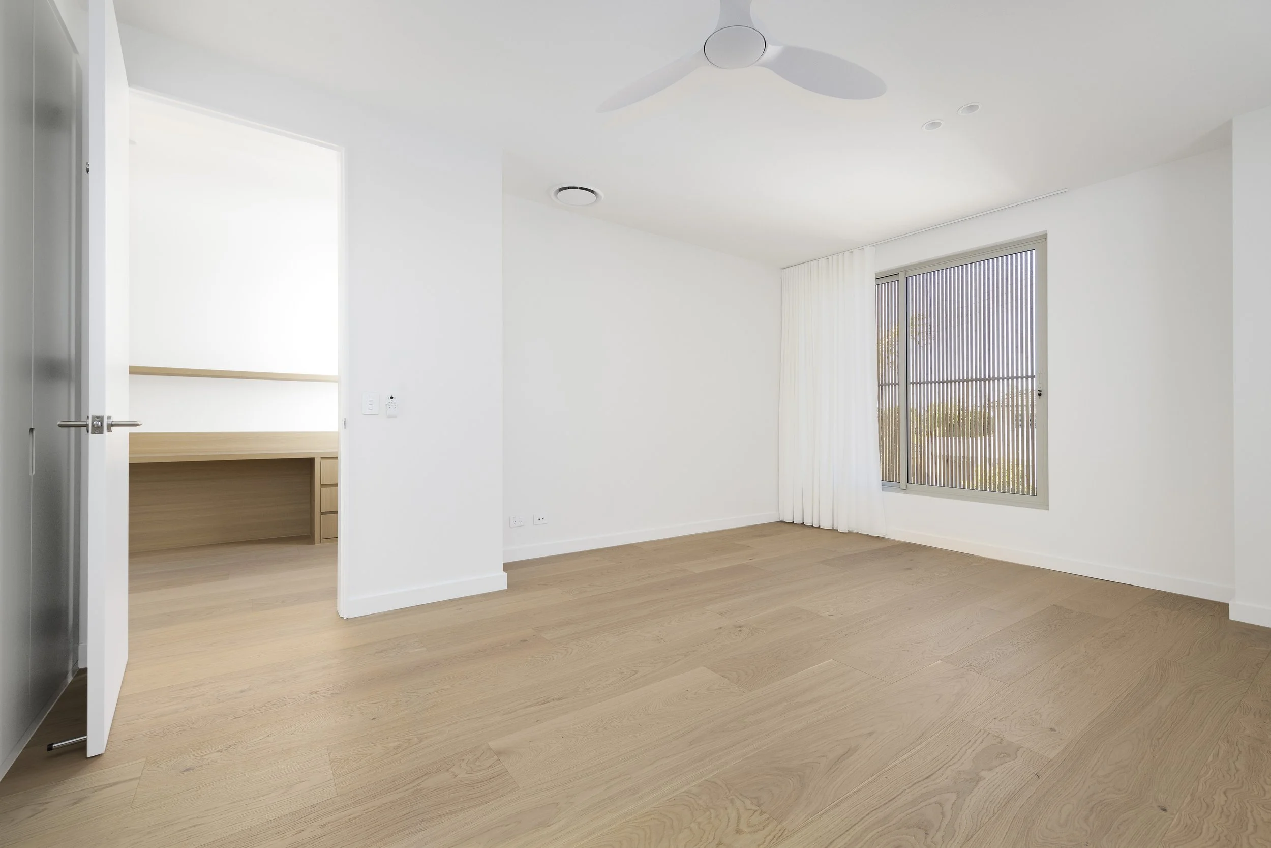 Empty room with wooden flooring, large window with vertical blinds, white curtains, and a ceiling fan. An open door leads to another room with a built-in desk or shelf.