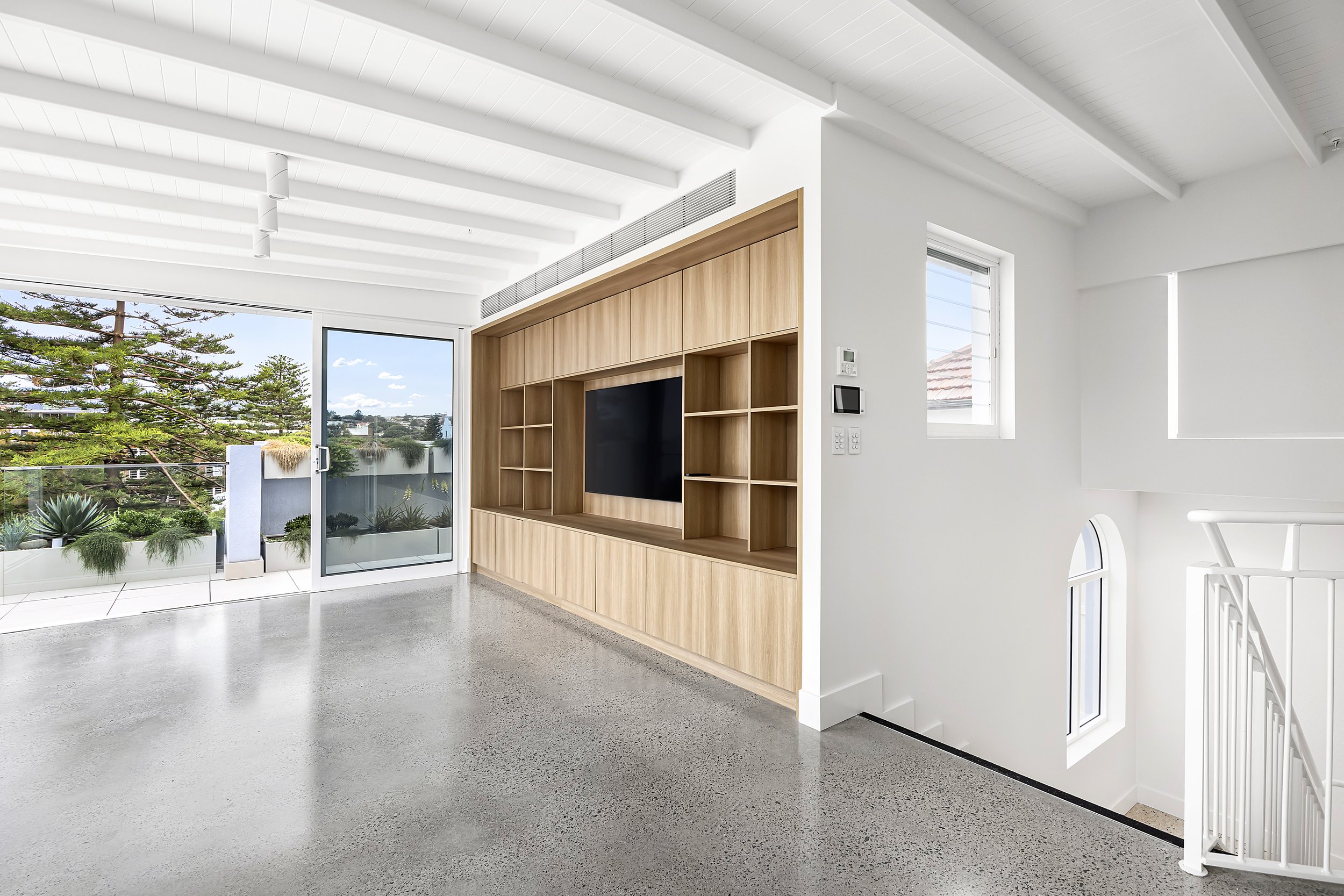 Modern living room with white walls, a wooden media wall with a TV, large glass sliding door leading to an outdoor balcony, and a staircase with a white railing.