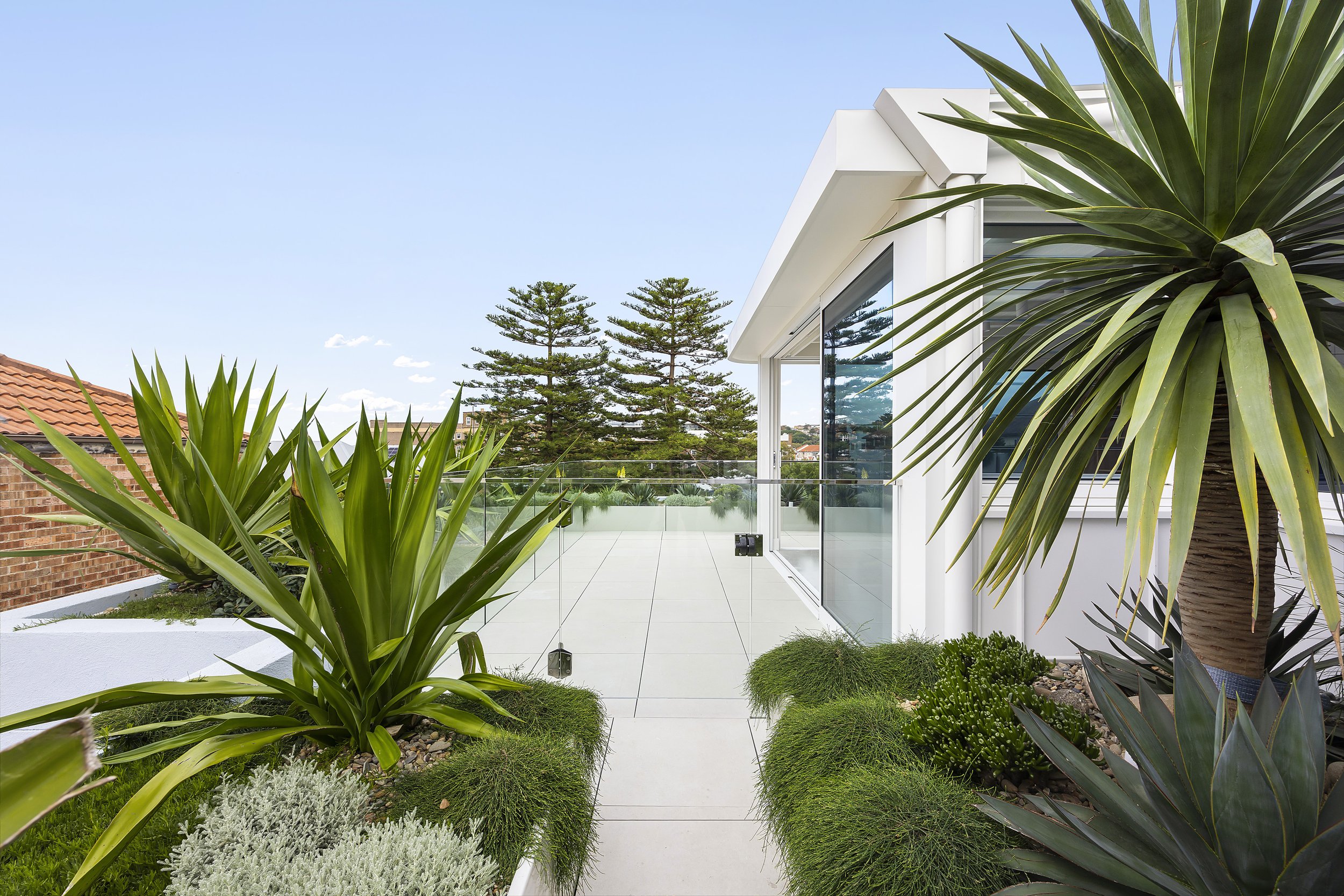 Modern balcony with glass railing, surrounded by green plants and trees, and a view of a blue sky.