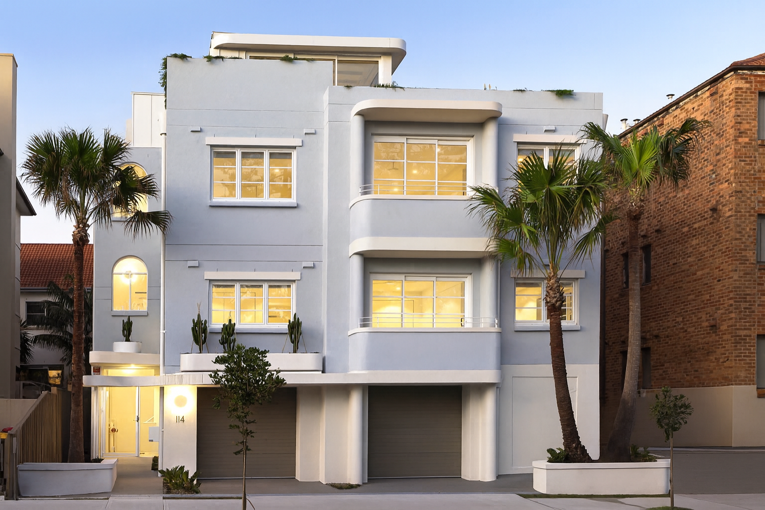 Modern white multi-story apartment building with rounded balconies, palm trees, and illuminated windows during dusk.